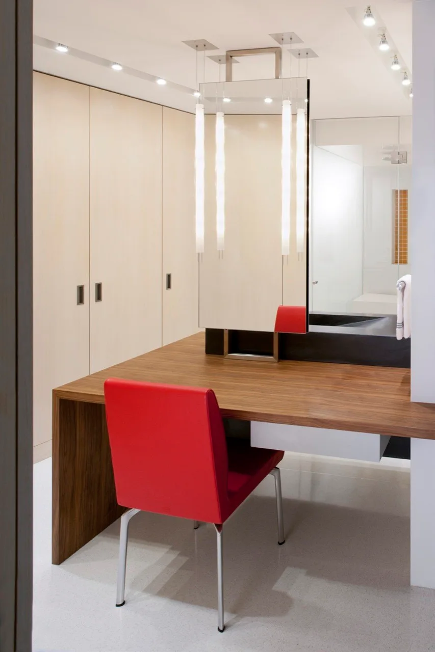 A minimalist dressing room features a warm wood desk with a single red chair positioned in front of floor-to-ceiling cream cabinetry.