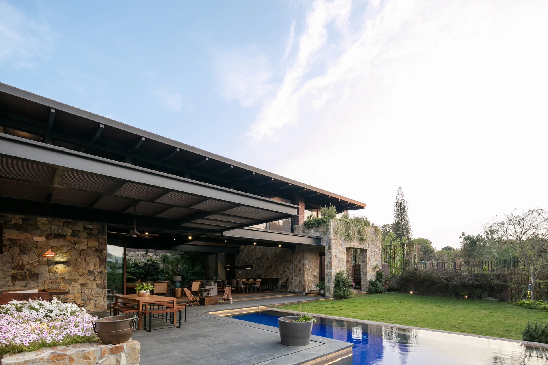 A modern covered patio with a stone wall features outdoor dining furniture next to a swimming pool and green lawn under a clear sky.