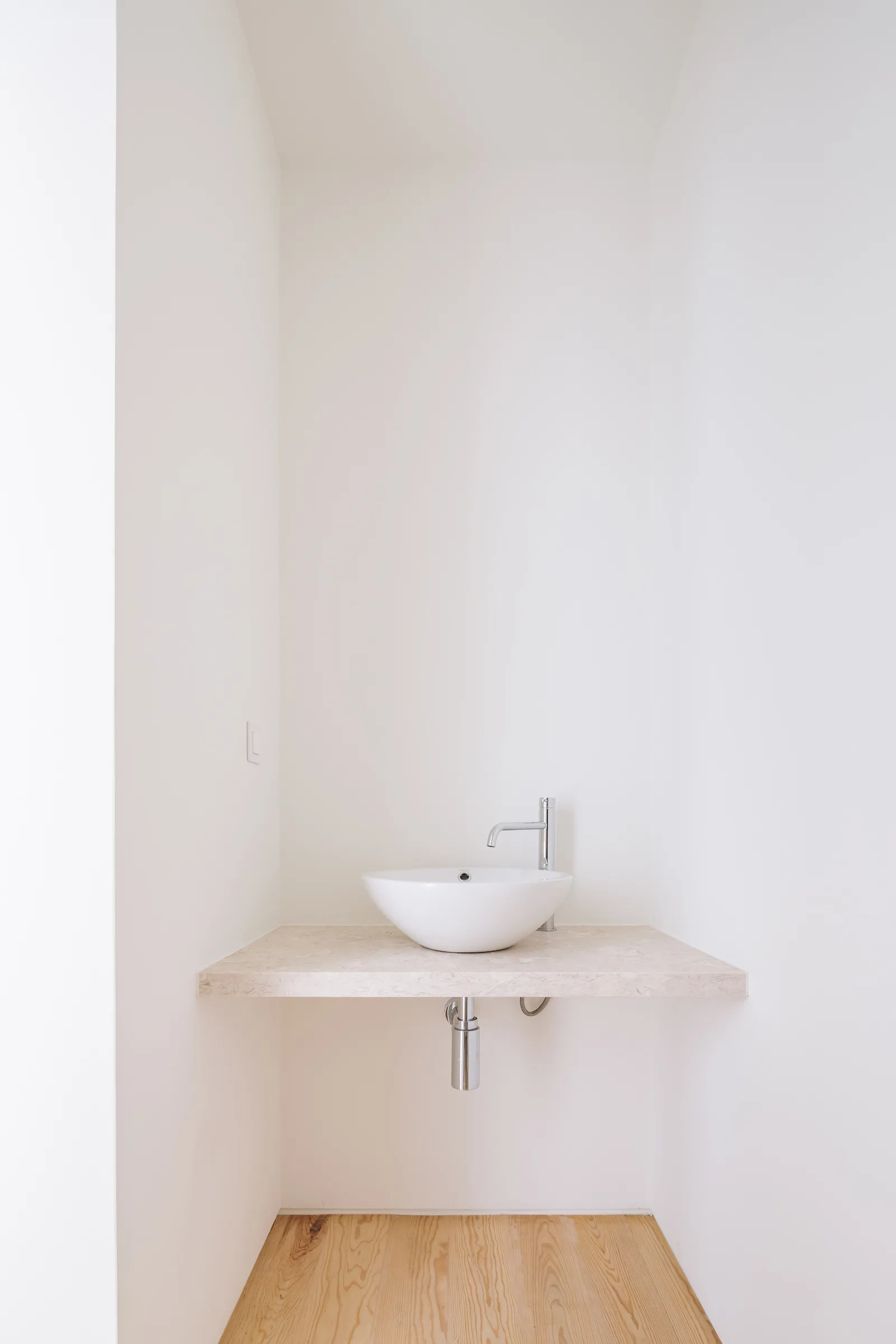 A minimalist white vessel sink rests on a floating beige stone vanity shelf inside a recessed wall niche above light wood floors.
