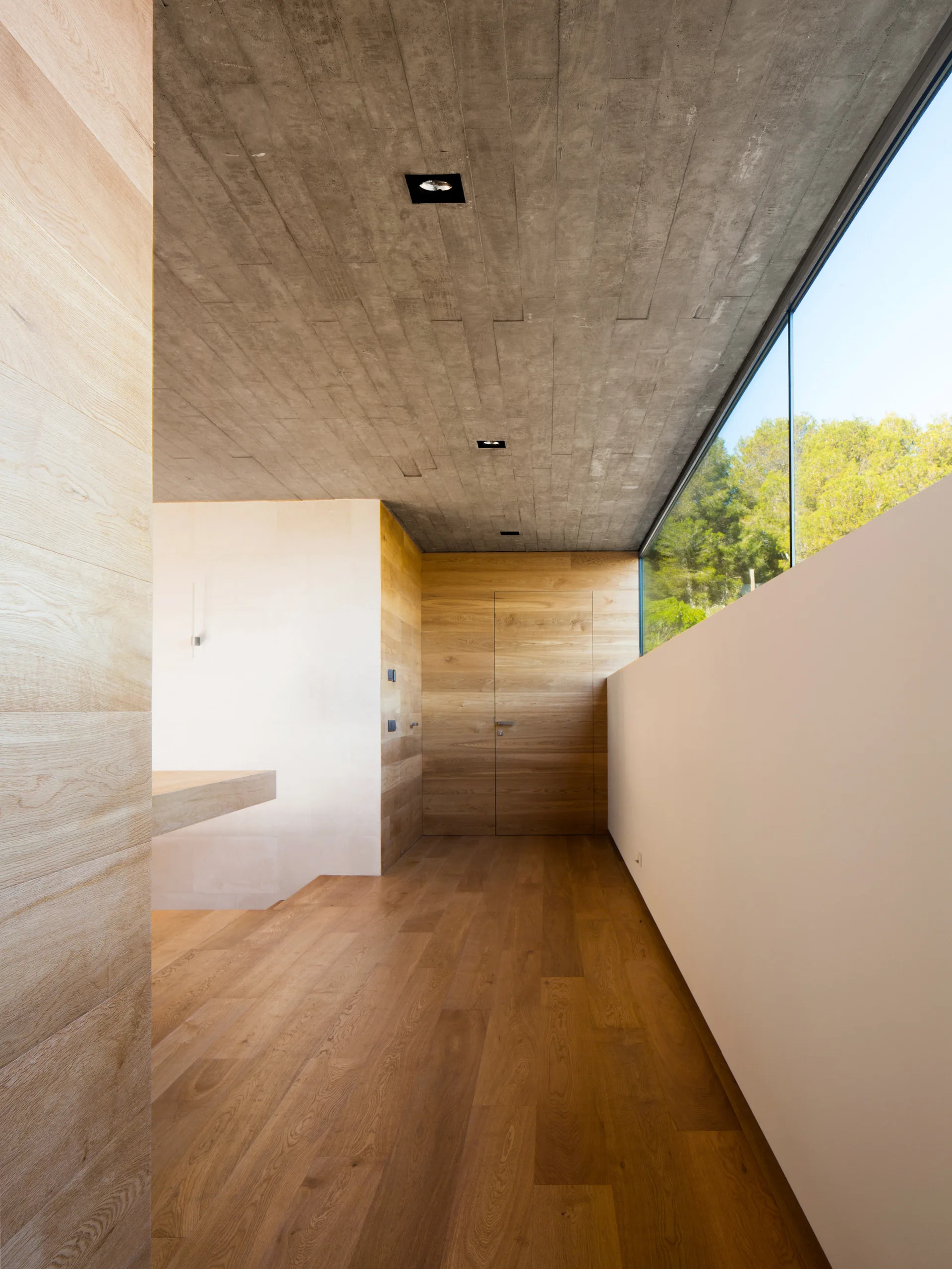 A minimalist hallway features natural wood flooring and wall paneling beneath a textured grey concrete ceiling with a long window showcasing green trees.