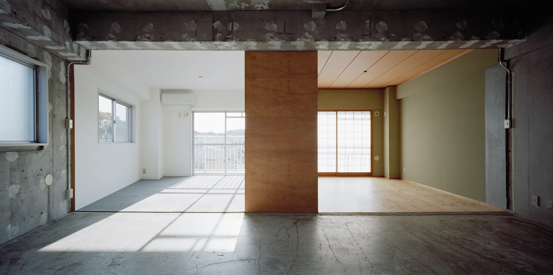 A minimalist apartment interior features an unfinished concrete floor, a central plywood partition wall, and a shoji screen window on the right.
