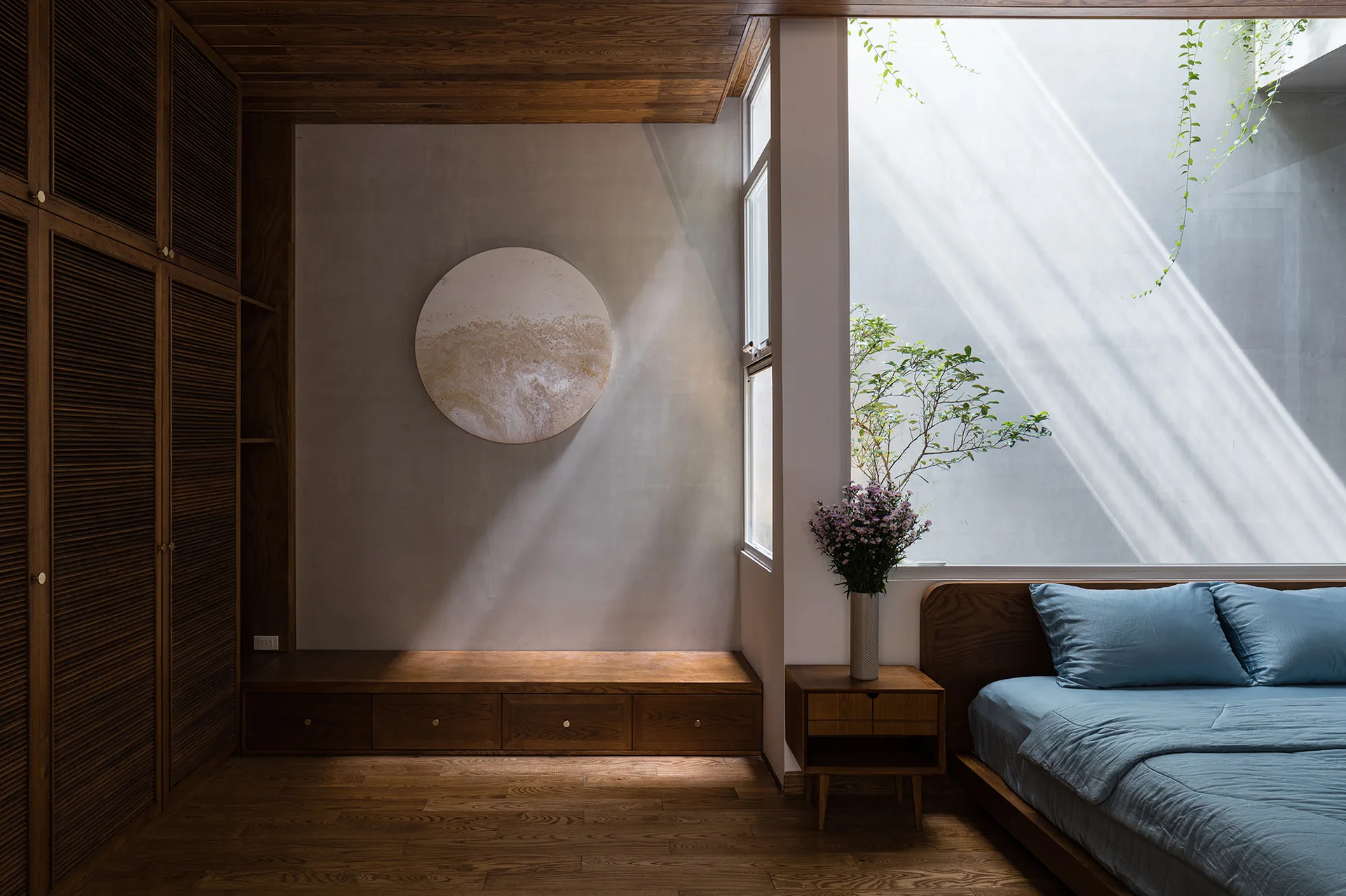 A minimalist bedroom with a low wooden bed, blue bedding, built-in louvered wood wardrobes, and a concrete wall illuminated by a tall light well.
