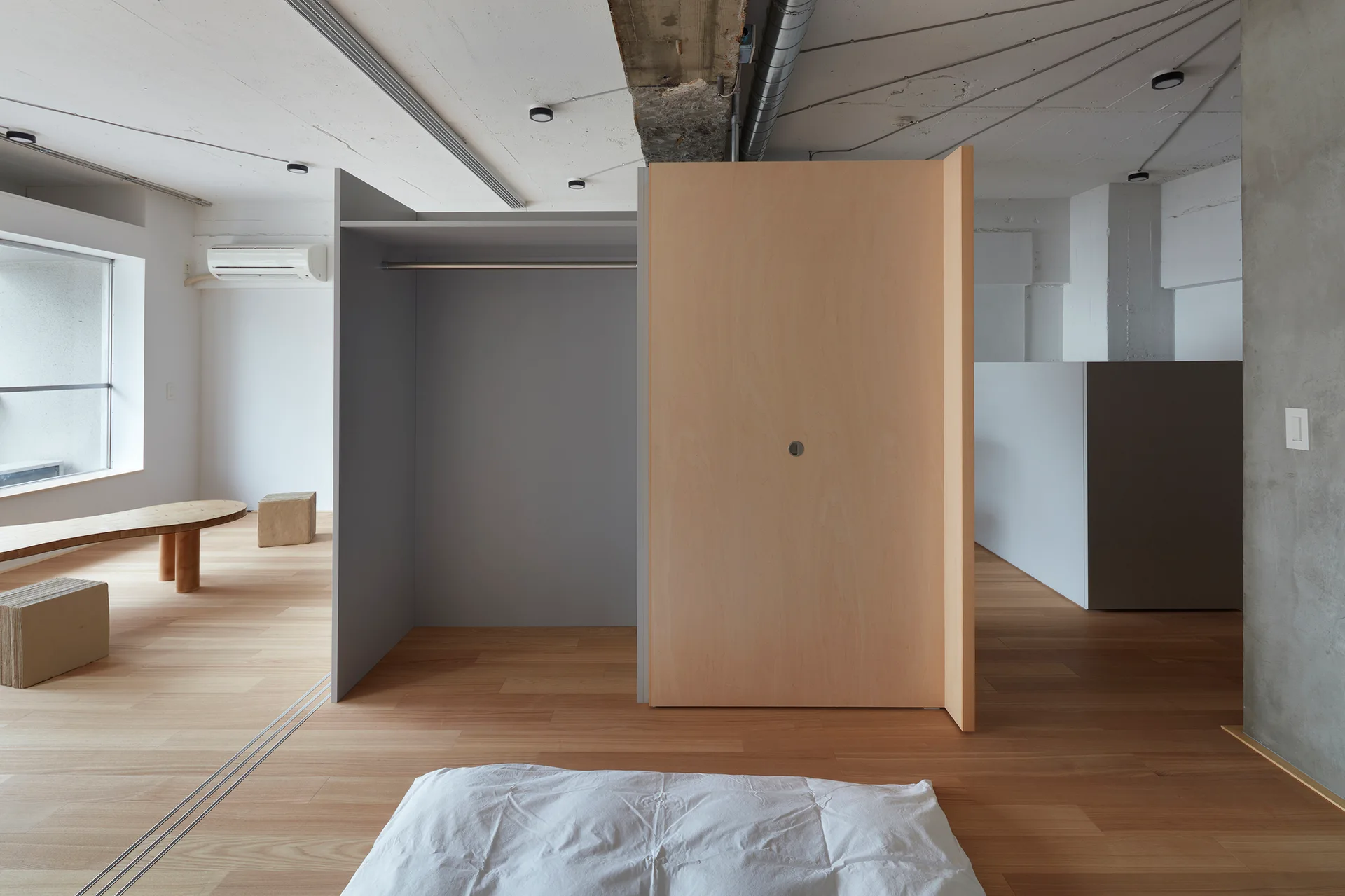 A minimalist bedroom features a futon on wood floors, a freestanding birch plywood partition closet, and an exposed concrete ceiling.