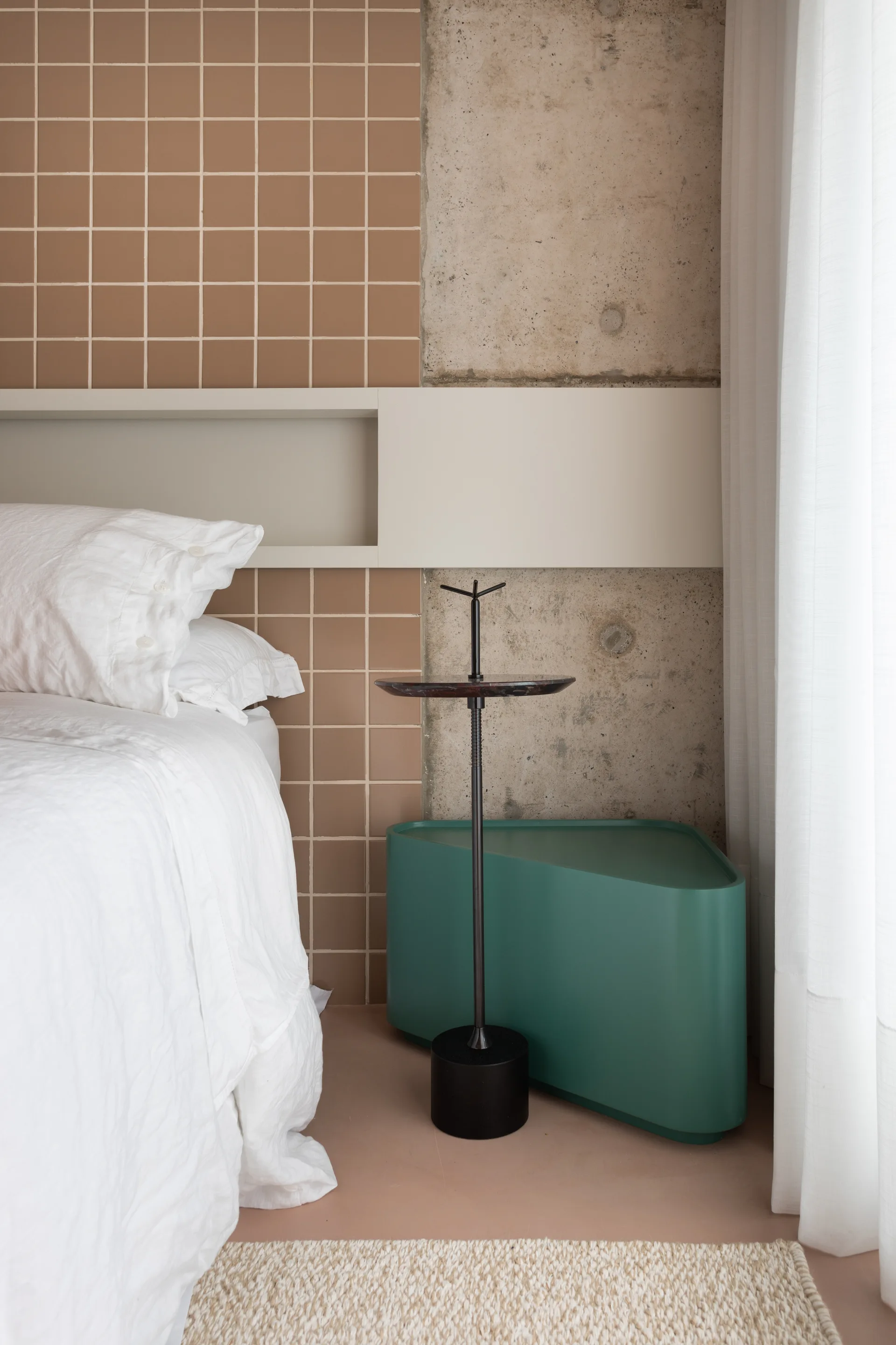 A bedside area with a white bed, a green triangular storage unit, a slim metal pedestal table, and a wall of terracotta tiles and raw concrete.