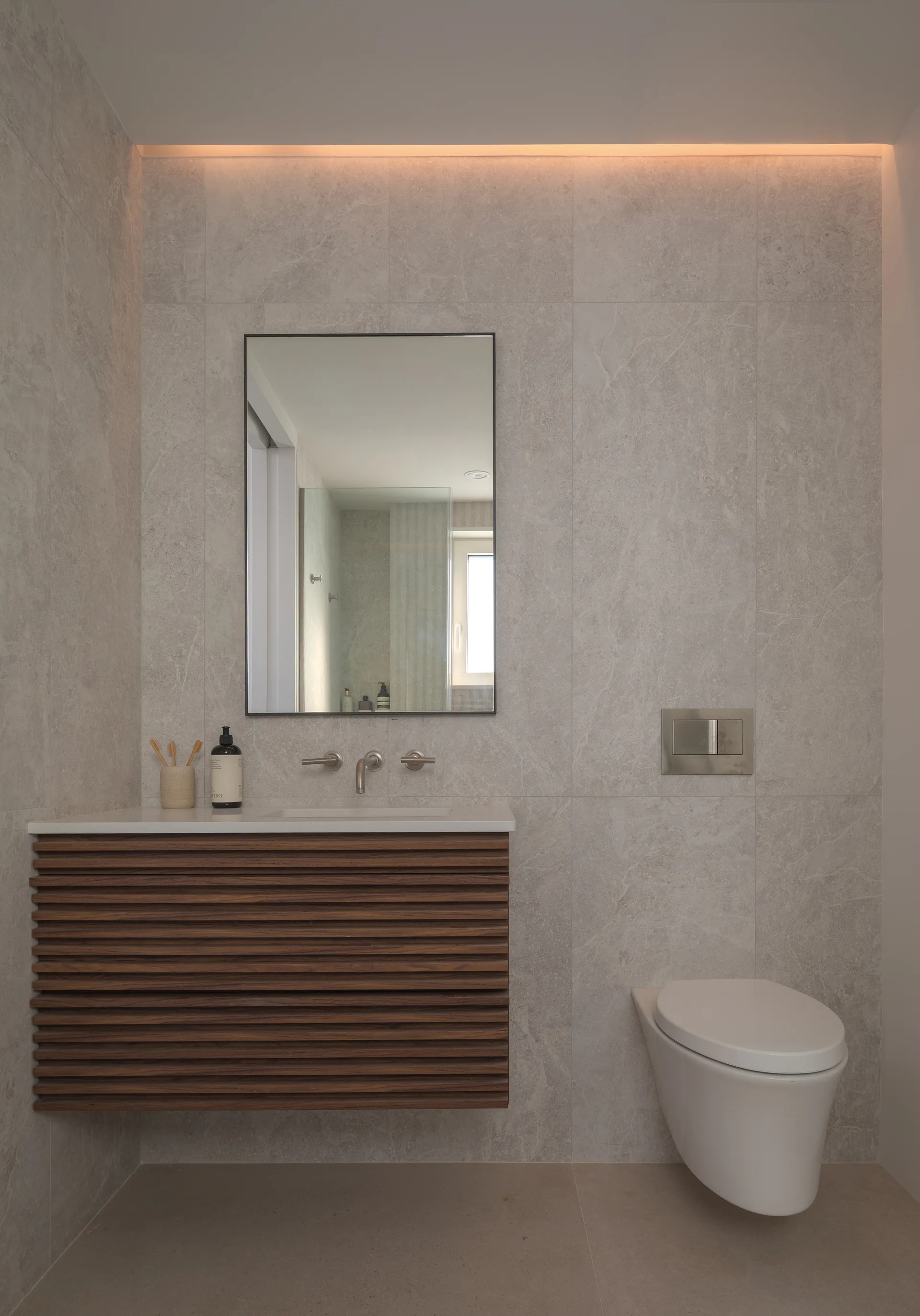 A modern bathroom features a wall-mounted dark wood slatted vanity and a white toilet against grey veined stone wall tiles under warm cove lighting.