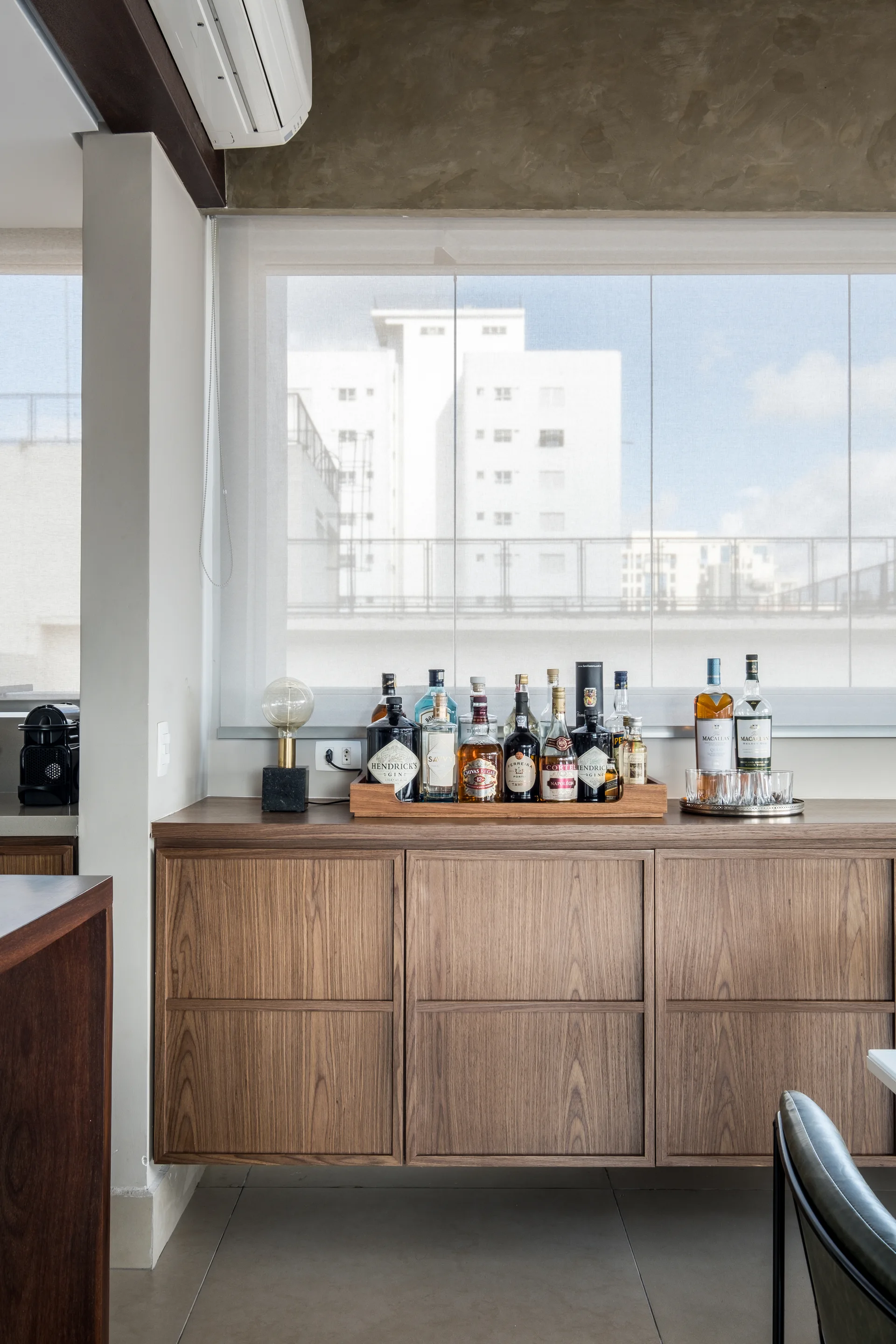 A modern home bar features a wall-mounted wooden cabinet with a collection of bottles and glassware set below a large window.