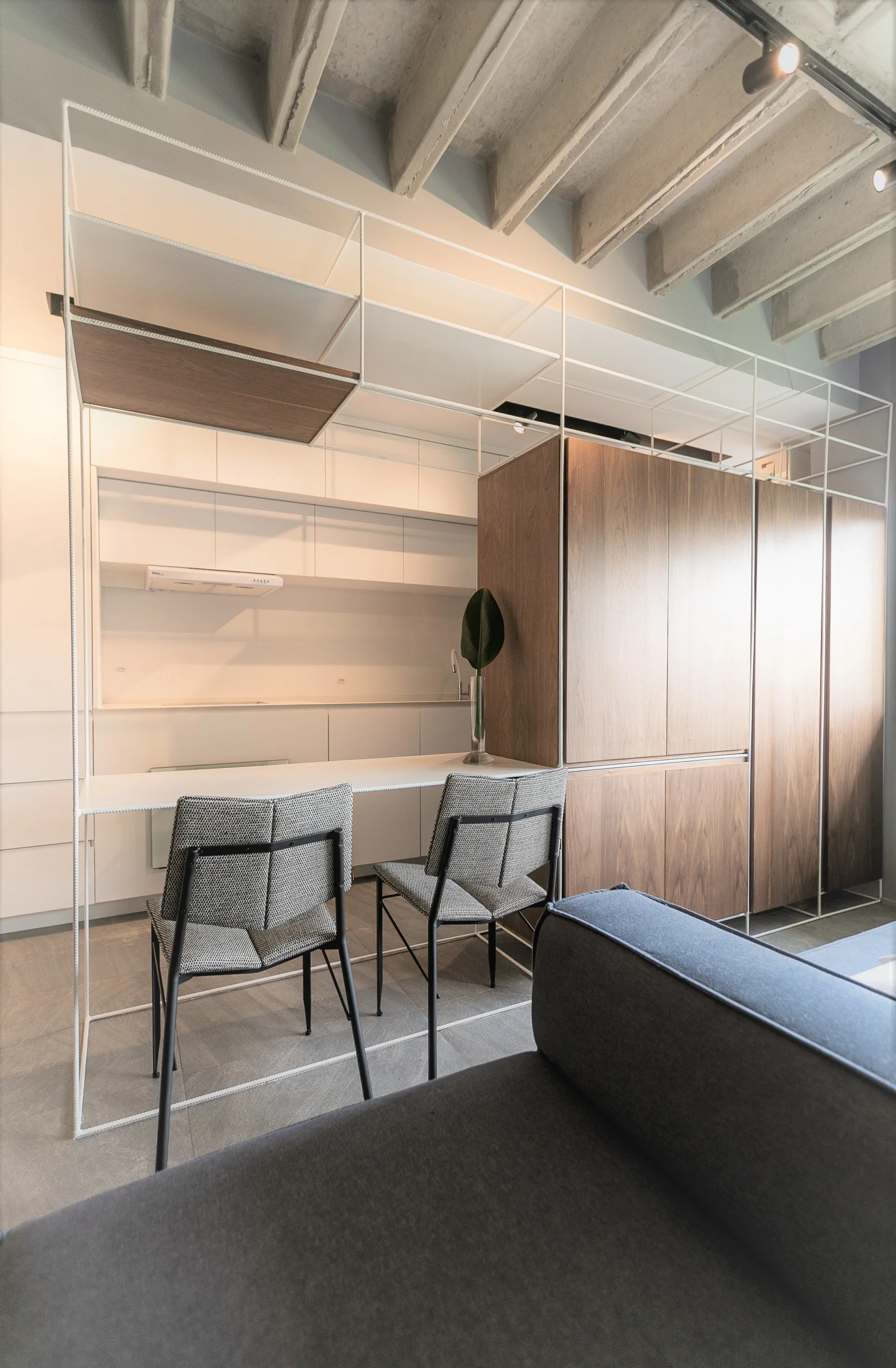 A modern kitchen featuring a white metal grid shelving unit enclosing a dining table, wood cabinetry, and an exposed concrete beam ceiling.
