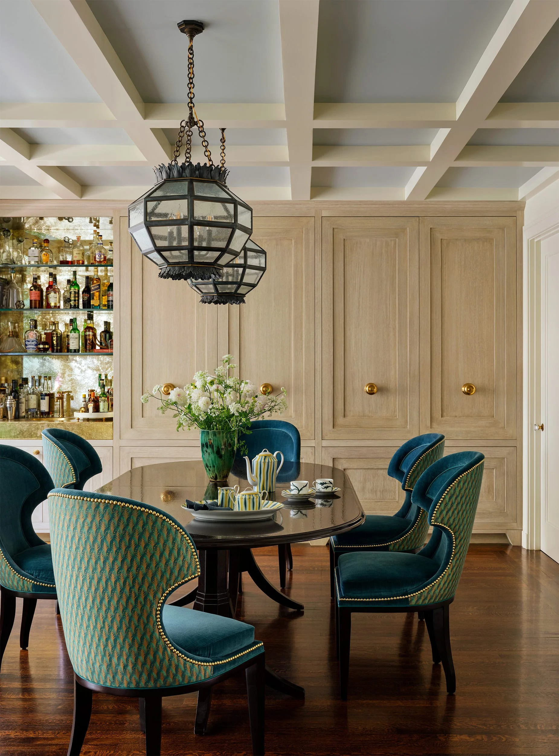 A formal dining room features a dark wood table, teal velvet chairs with gold nailhead trim, a coffered ceiling, and light wood built-in cabinets.