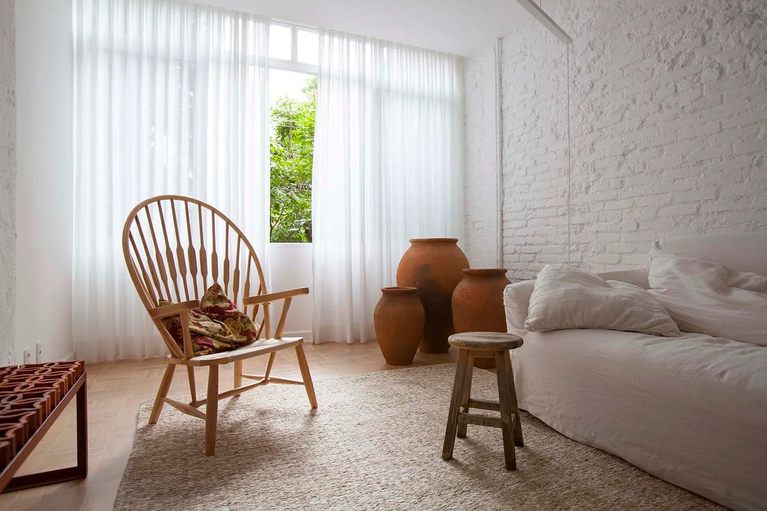 A light-filled living room featuring a white sofa, a wooden spindle chair, a small stool, and three large terracotta vases against a painted brick wall.