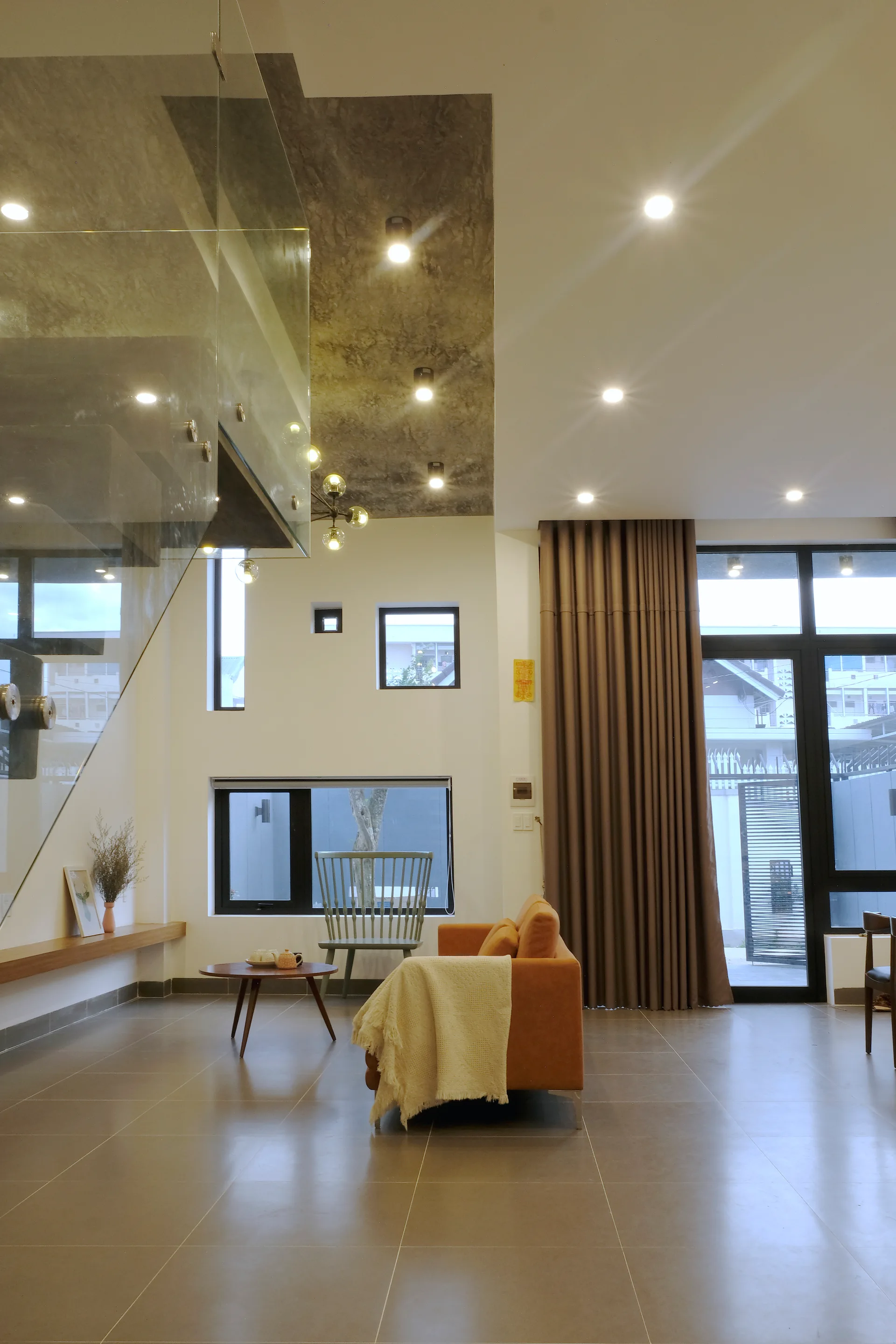 A modern living room featuring an orange sofa with a beige throw, a glass-railed staircase, and a gray concrete-textured ceiling section.