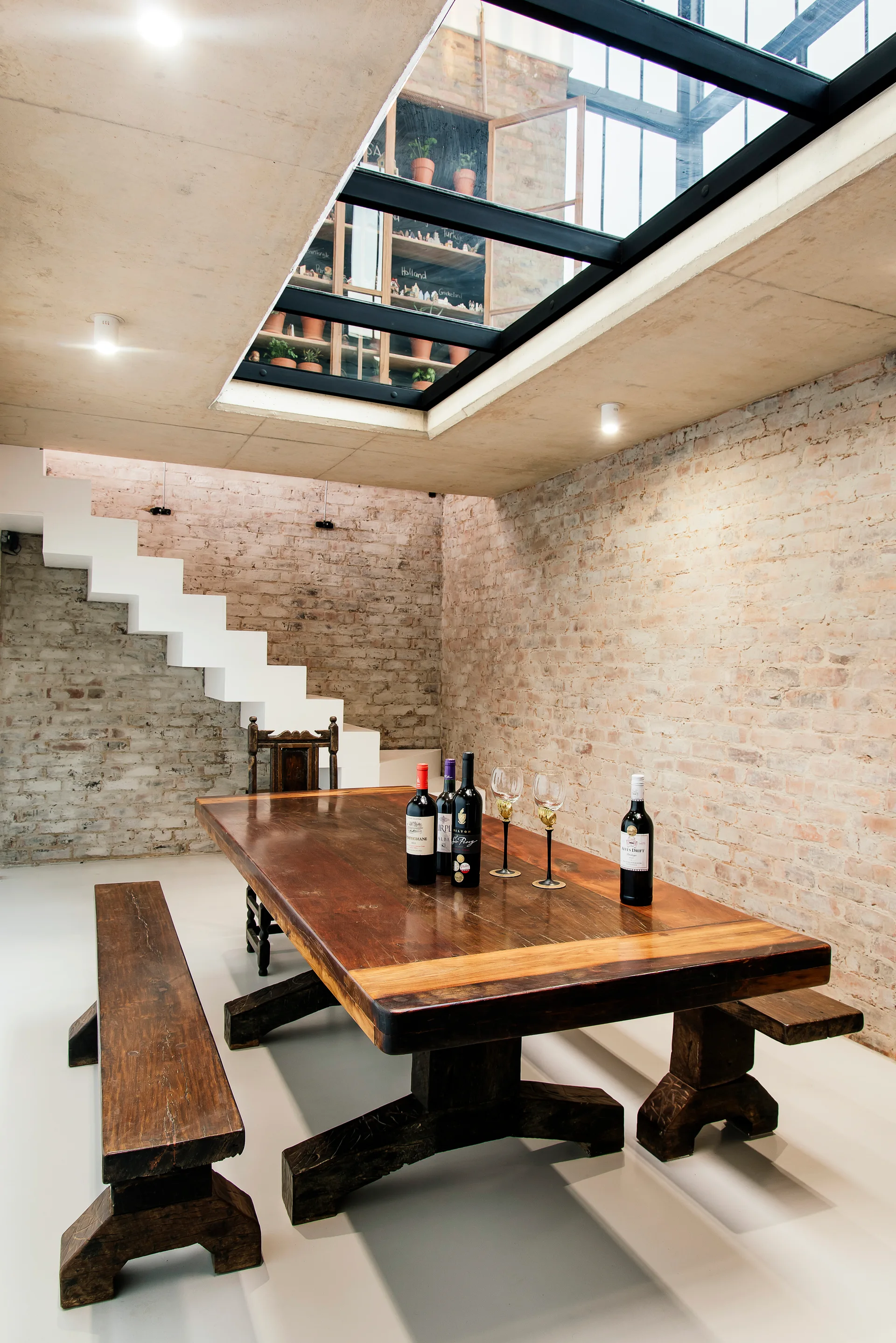 A rustic dining table and benches sit below a large glass skylight in a room with exposed brick walls and a white staircase.