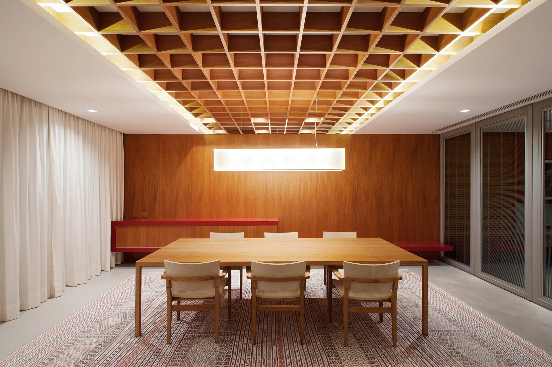 A modern dining room features a large wood table, matching chairs on a patterned rug, a wood-paneled wall, and an illuminated coffered ceiling.