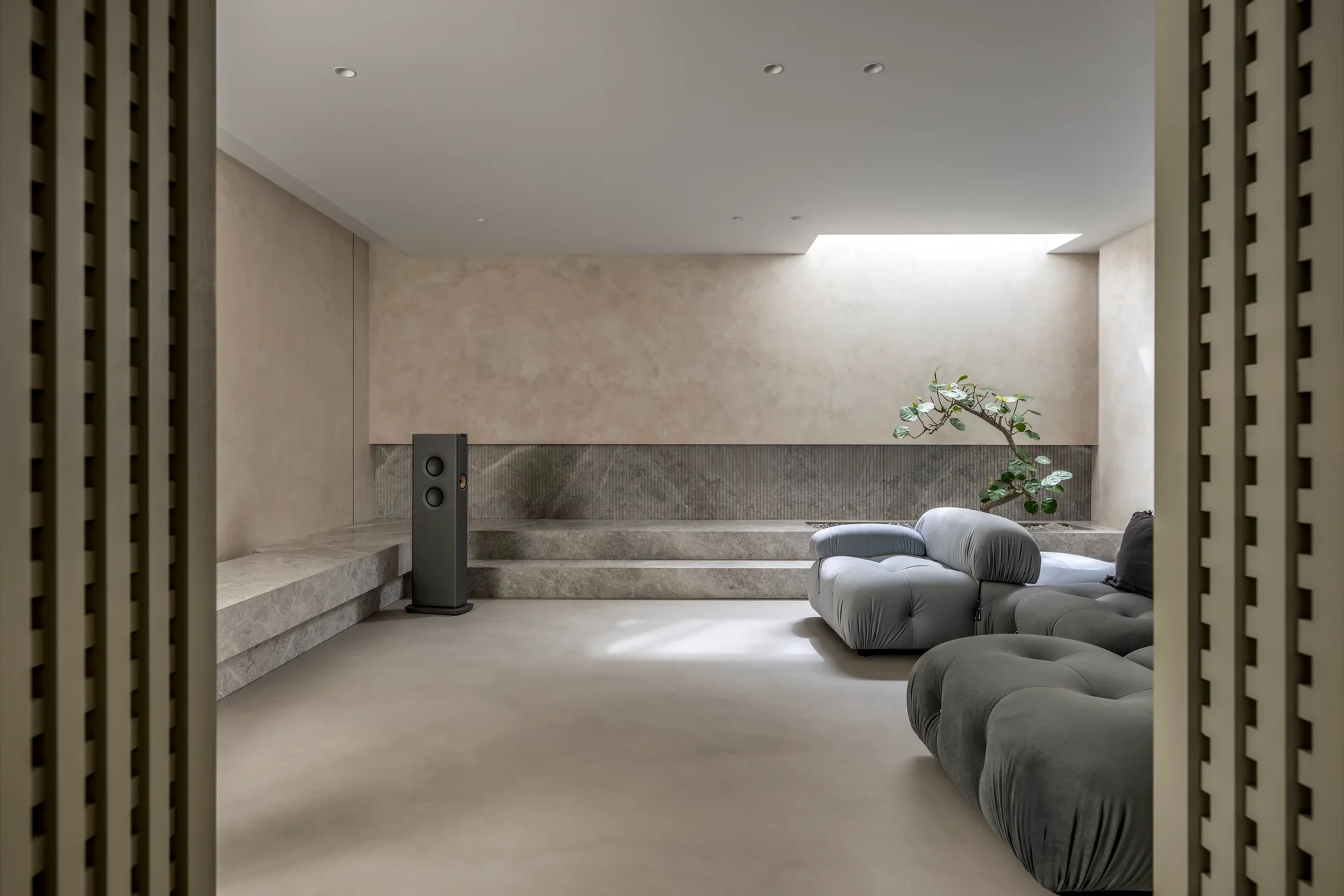 A minimalist grey and beige living room features a tufted sectional, a stepped stone ledge, and a ceiling skylight illuminating a small tree.