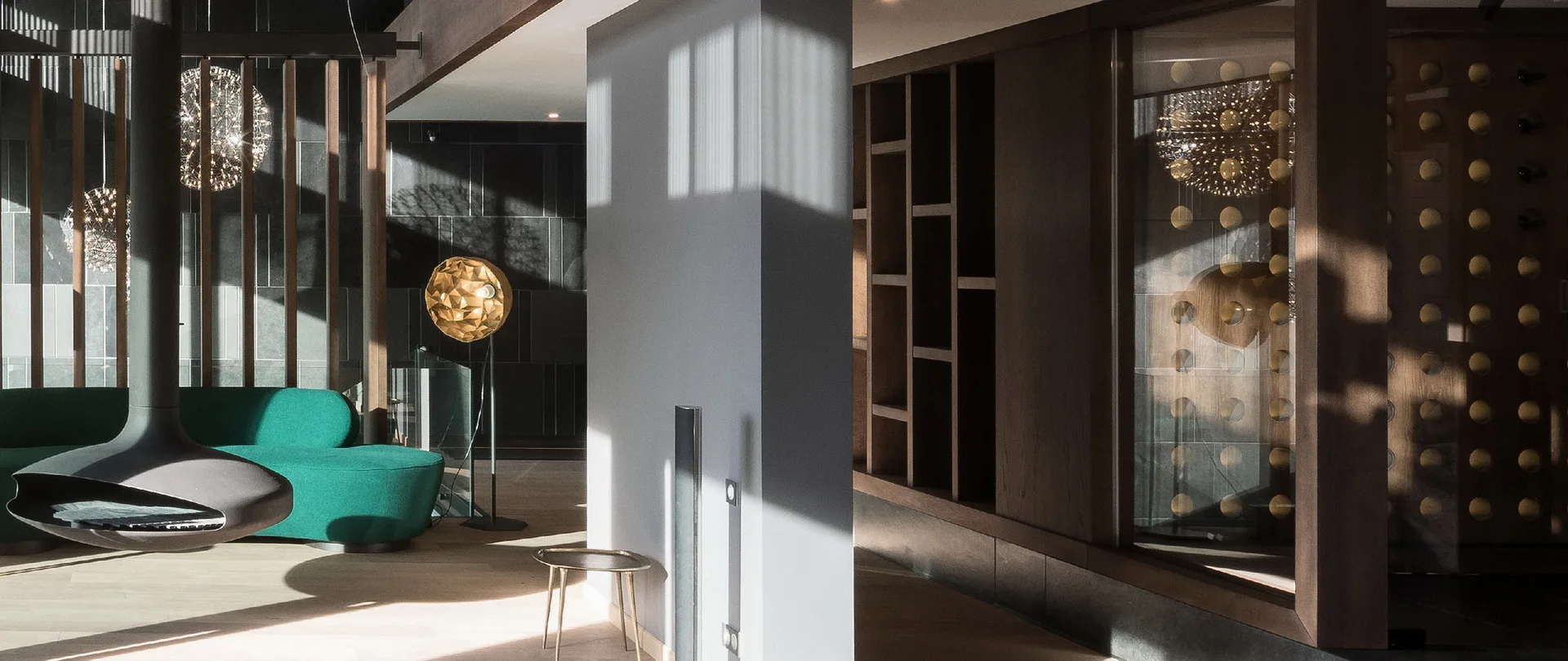 A modern lounge featuring a curved emerald green sofa near a black suspended fireplace, separated from dark wood shelving by a grey pillar.