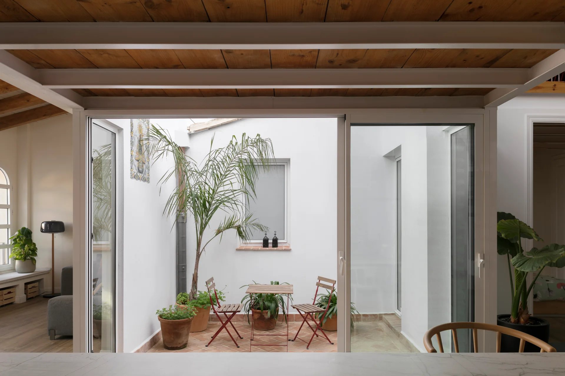 A bright white-walled courtyard with a small wood and red metal bistro set, terracotta potted plants, and tile flooring, viewed through glass doors.
