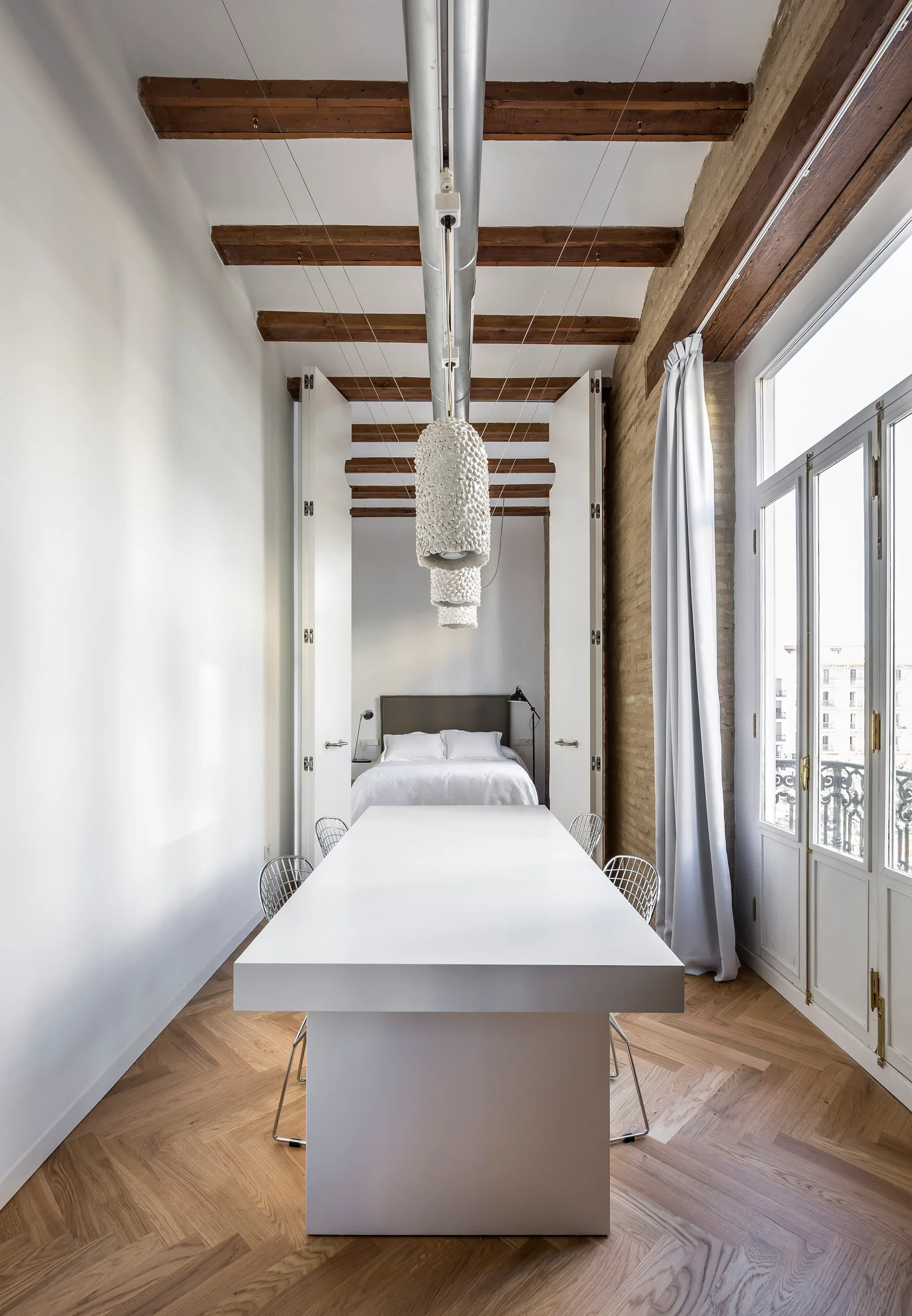 A long white dining table with wire chairs sits under exposed wooden beams in a loft featuring herringbone wood floors and a bedroom in the background.