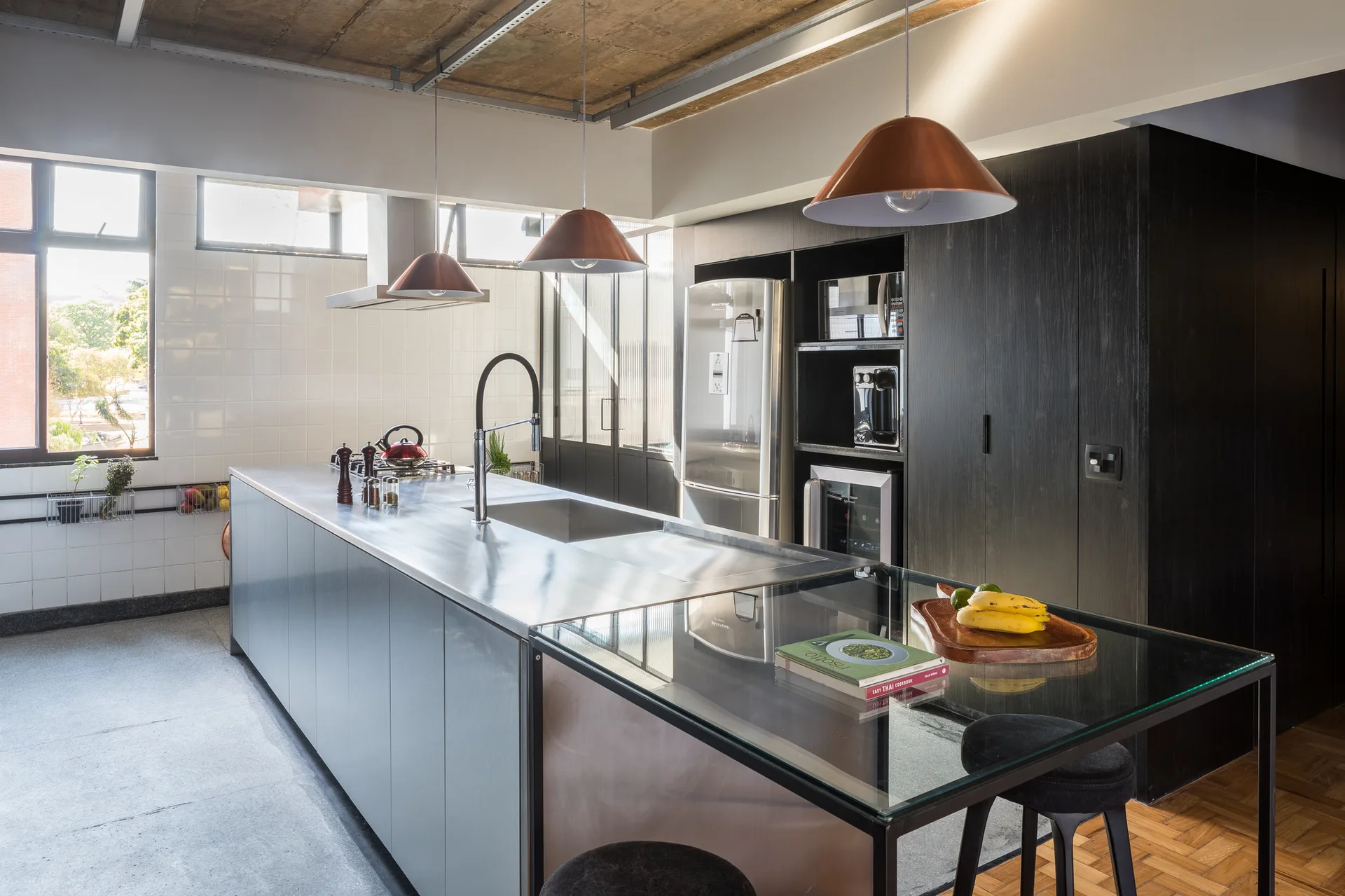 A modern kitchen features a stainless steel island with copper pendant lights, an exposed concrete ceiling, and a wall of matte black cabinets.