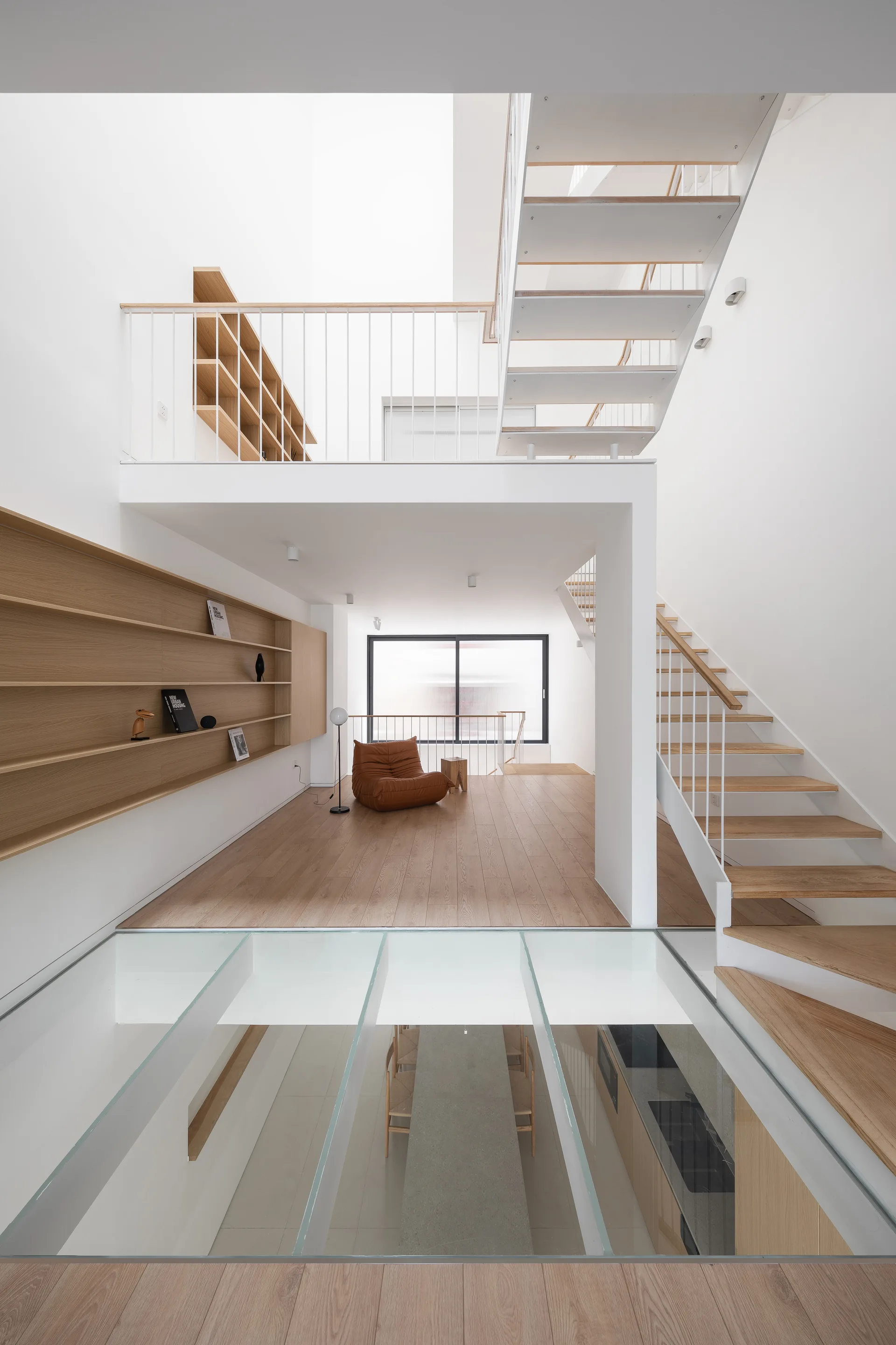 A modern multi-level interior features a white steel staircase, wooden floors, a glass floor panel viewing the level below, and built-in shelving.