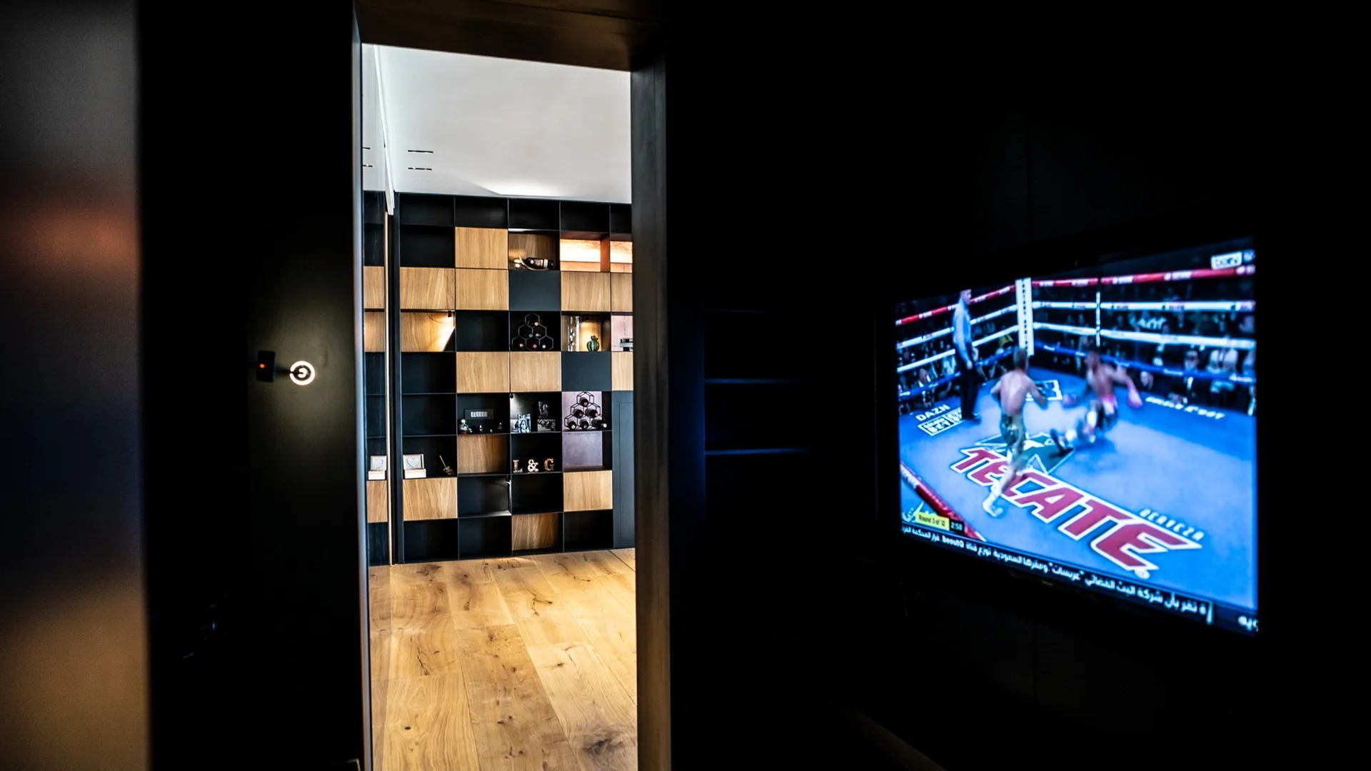 View through a dark doorway into a room with a black and wood checkerboard shelving unit, hardwood floors, and a television.