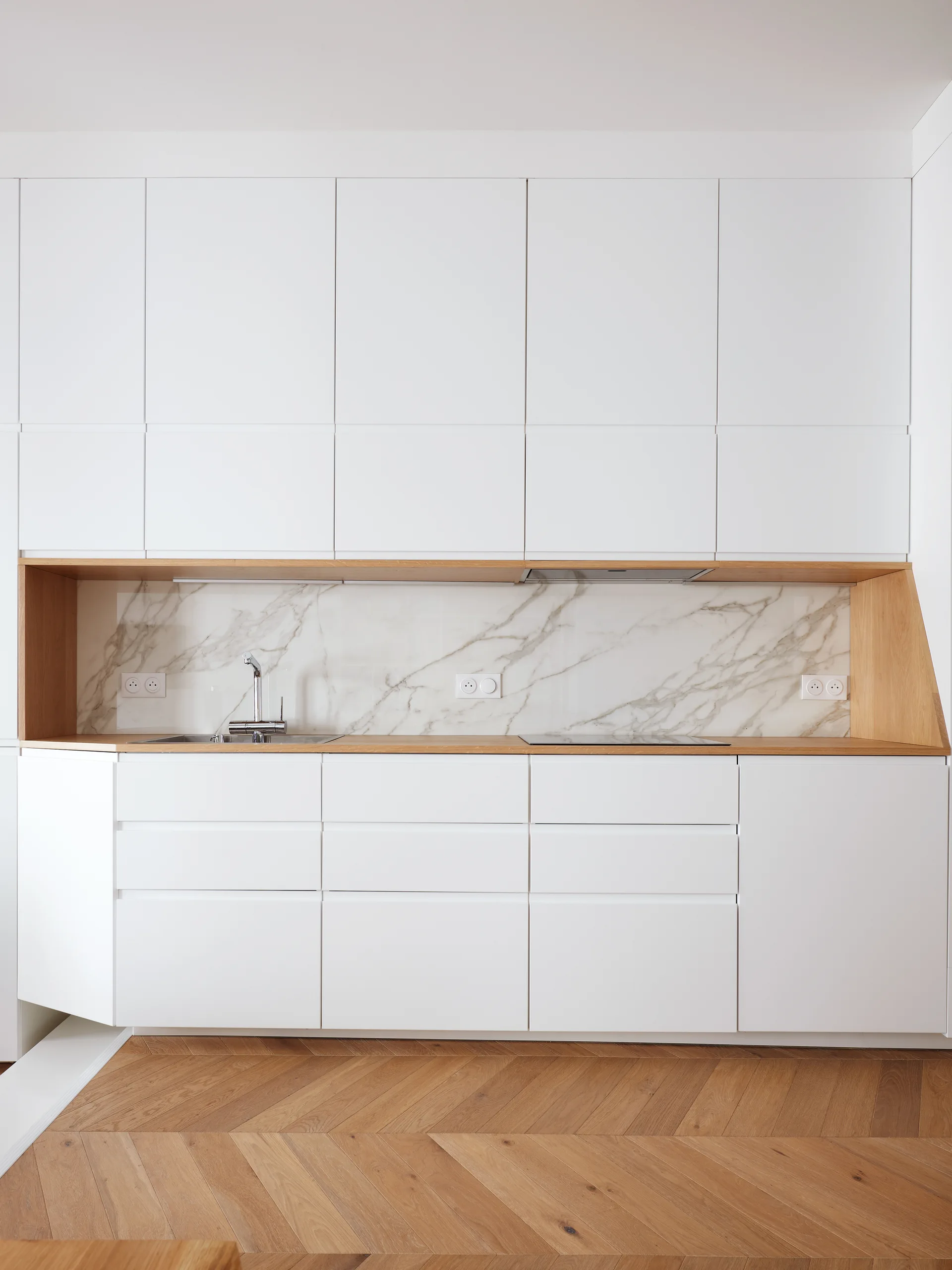 A modern minimalist kitchen features seamless white cabinets surrounding a wood-lined niche with a marble backsplash and herringbone wood floor.