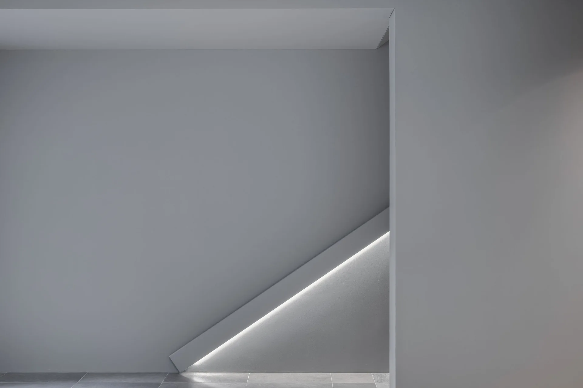 A minimalist grey staircase wall features a diagonal recessed architectural groove illuminated by a bright, integrated linear LED light strip.