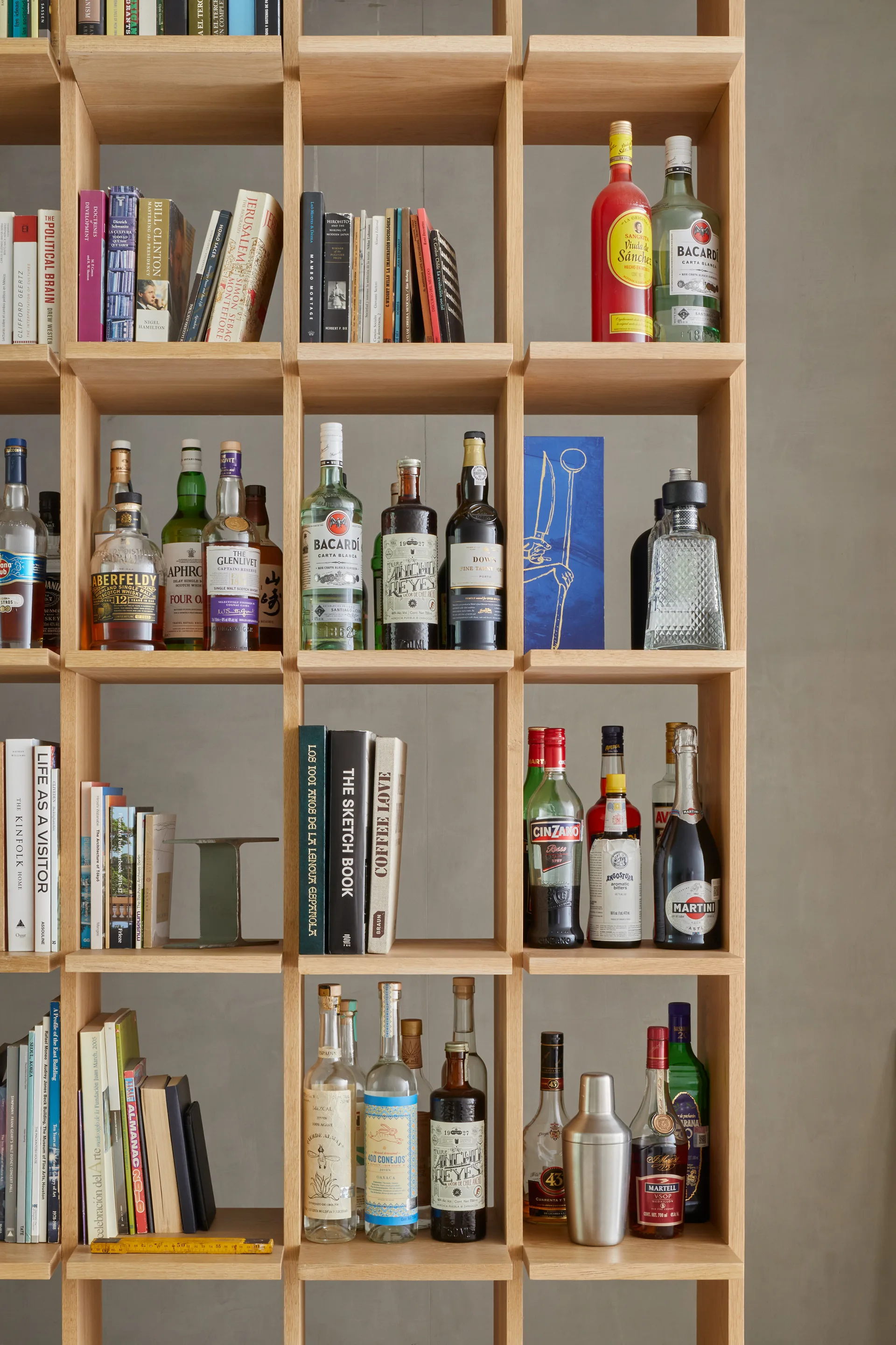 A light wood grid shelving unit filled with organized rows of books, various liquor bottles, a cocktail shaker, and a small blue art print.
