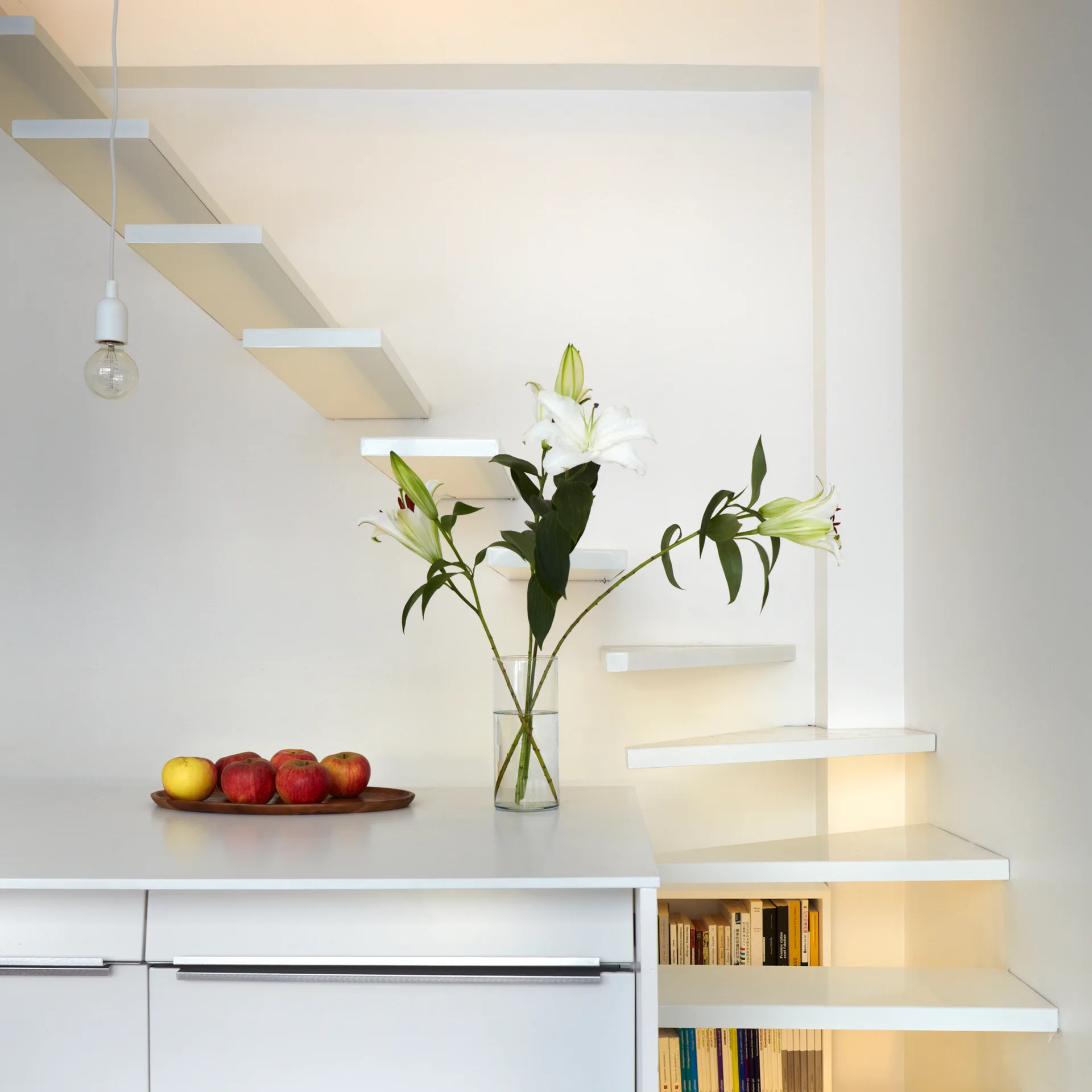 A white minimalist floating staircase ascends next to a white cabinet with a glass vase of lilies and a wooden tray of apples.