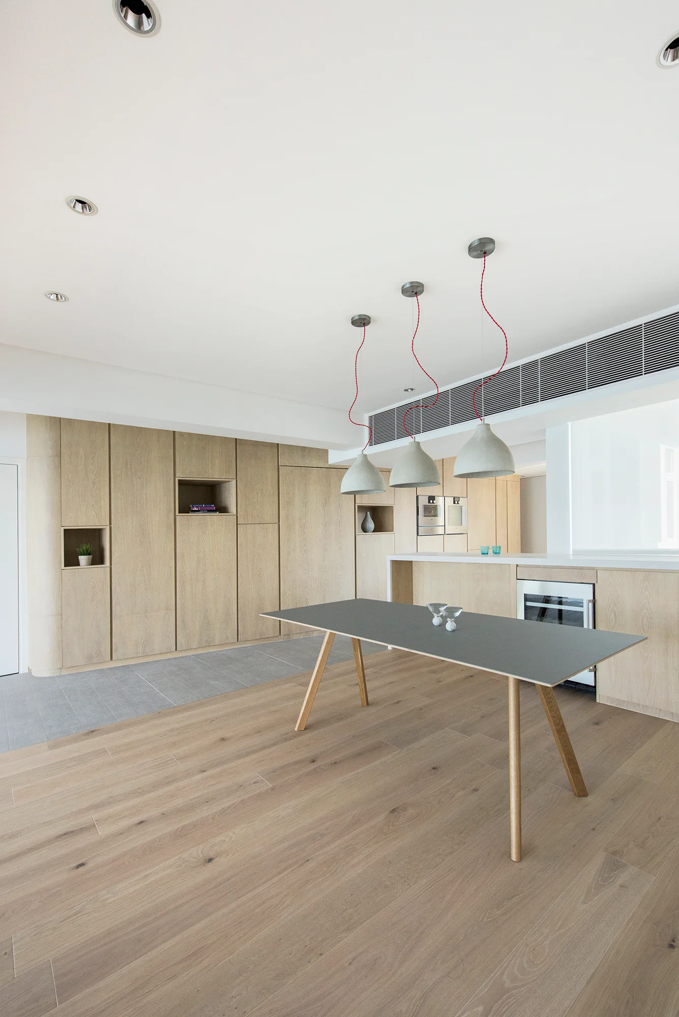 A modern kitchen featuring floor-to-ceiling light wood cabinetry with built-in niches, a grey dining table, and three pendant lights with red cords.