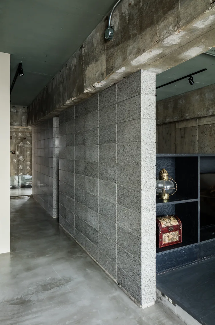 An industrial hallway with a textured cinder block partition wall, raw concrete beam, polished grey floor, and black open shelving.