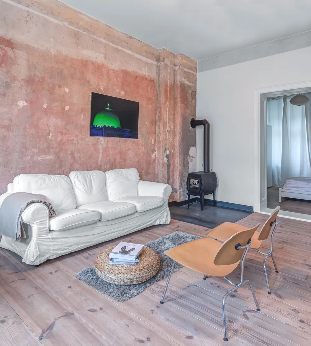 A white sofa sits against a distressed terracotta plaster wall in a living room with light wood floors, a black wood stove, and two wooden chairs.