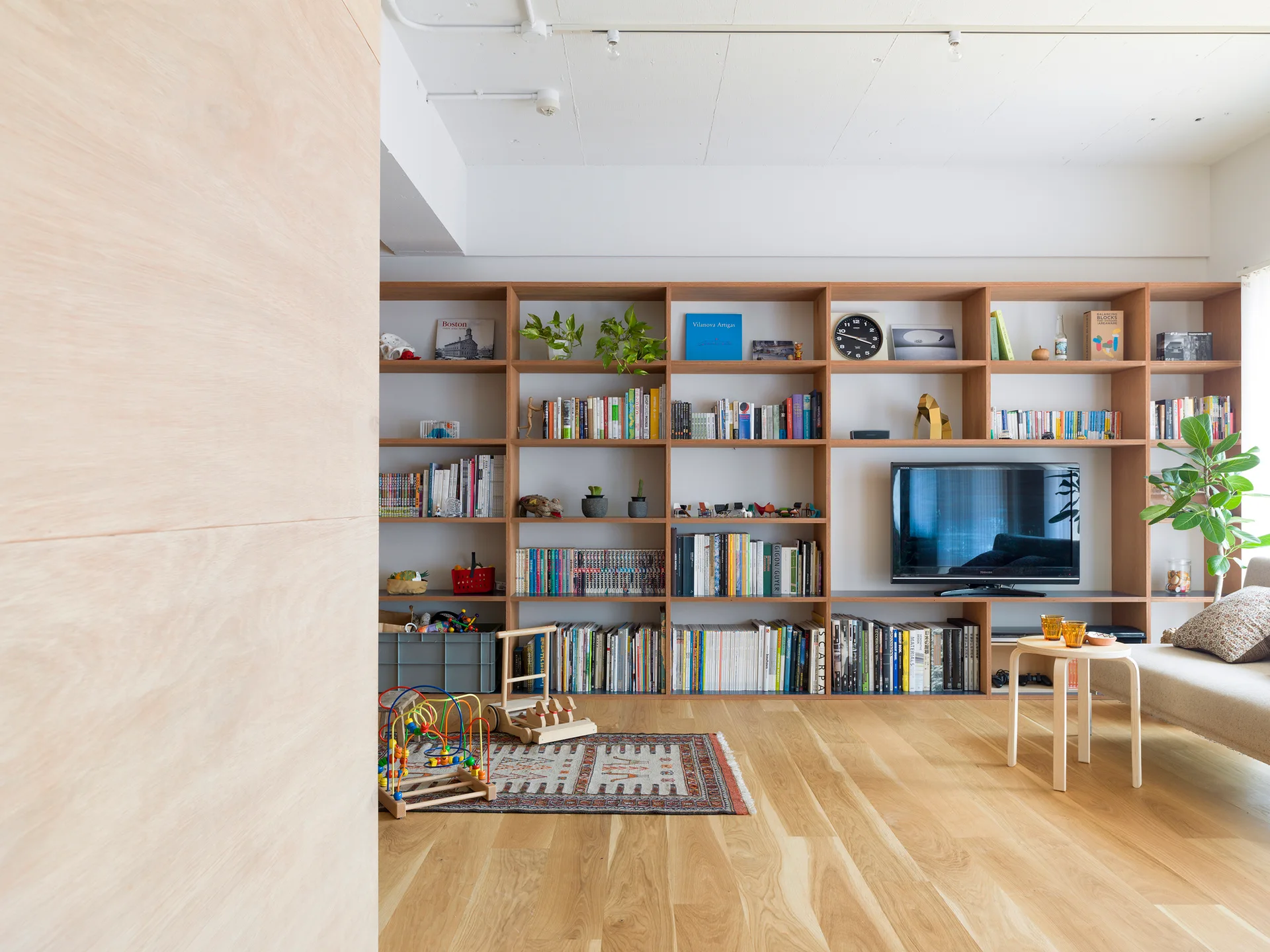 A bright living room features a floor-to-ceiling light wood grid bookshelf holding books, plants, and a television above a light wood floor.