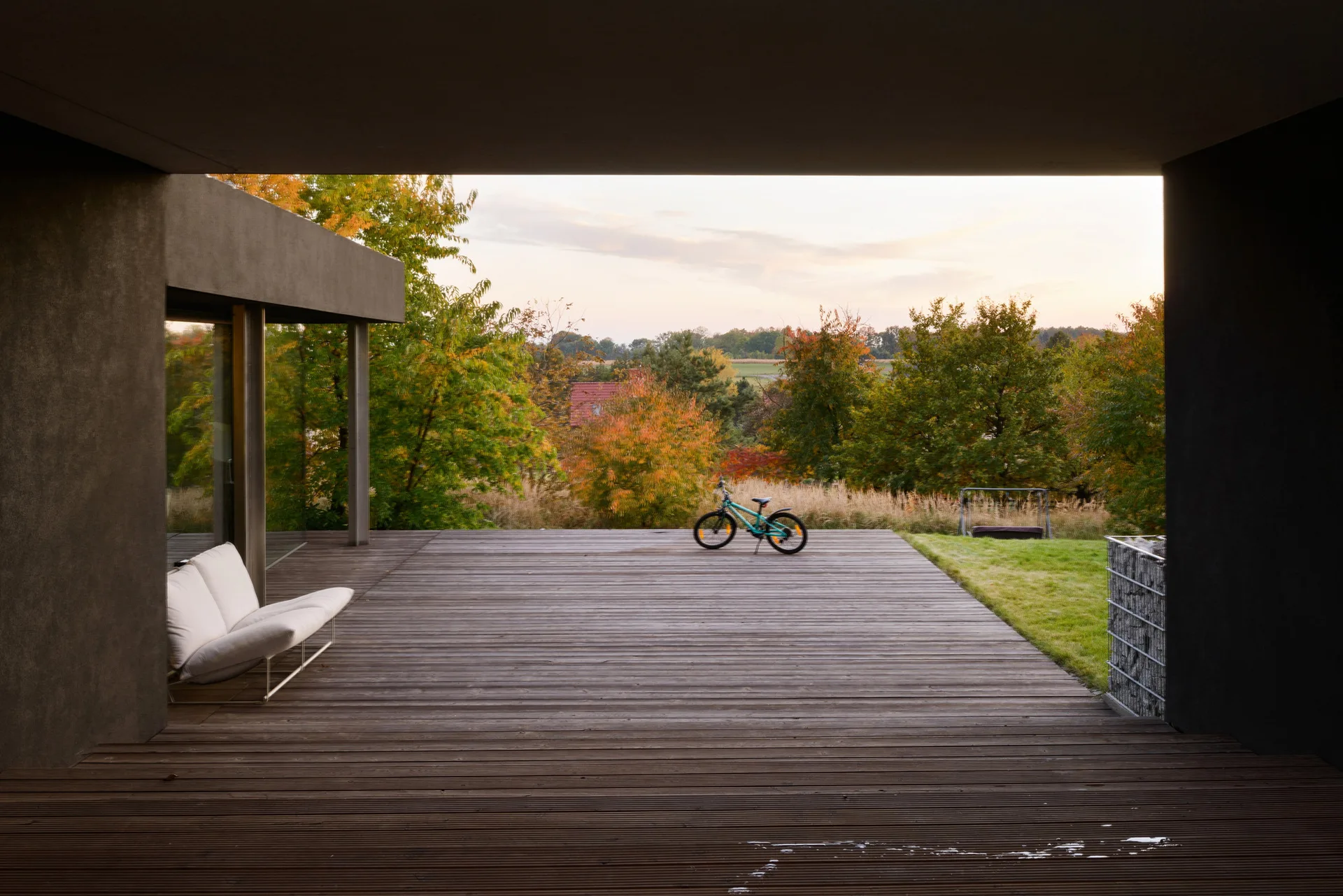 A minimalist covered wooden terrace features a white outdoor sofa, dark gray framing walls, and a view of an autumnal lawn and trees.