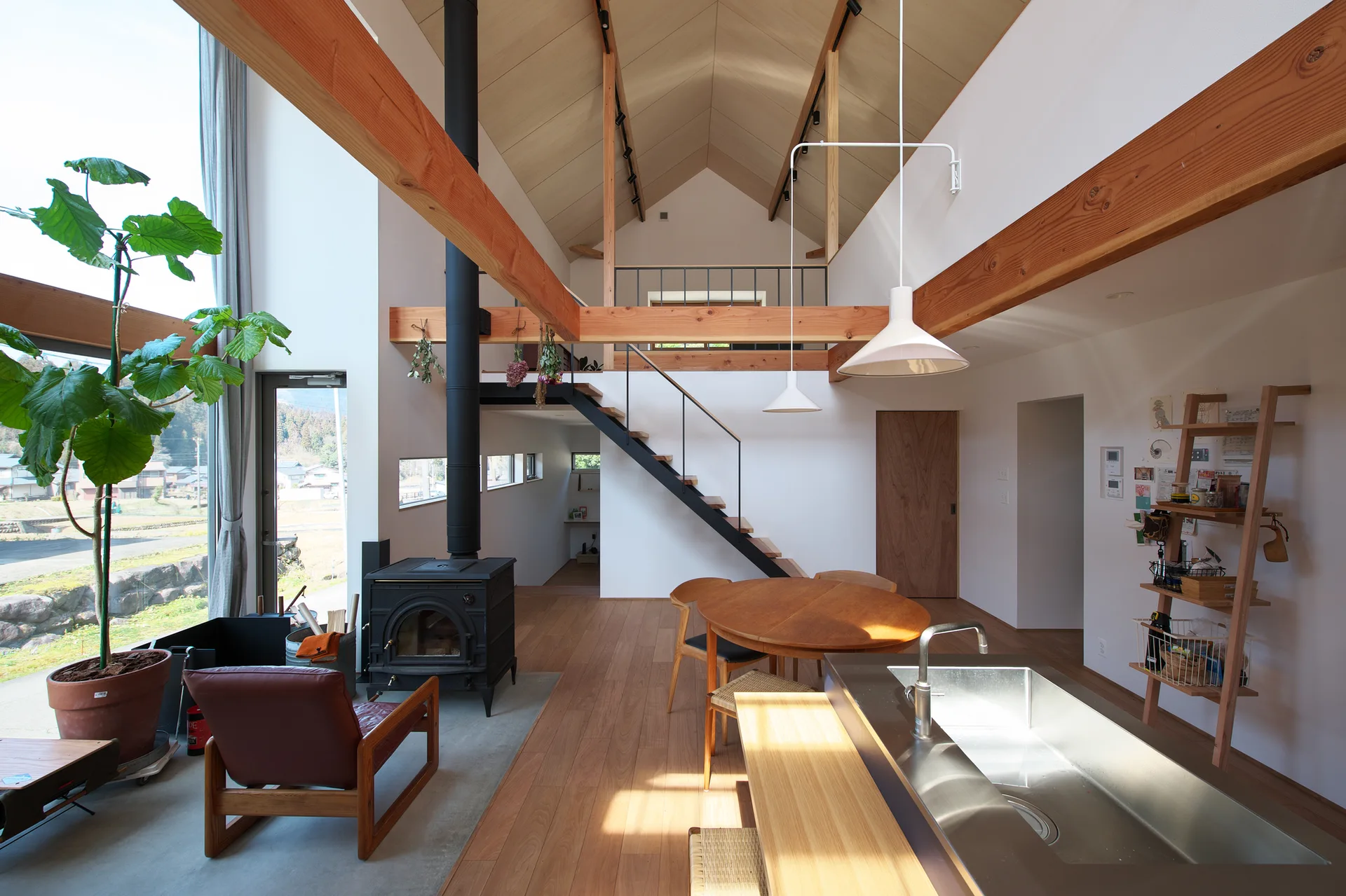 An open living space featuring a vaulted wood ceiling, exposed beams, a black wood-burning stove, a wooden dining table, and an open staircase.