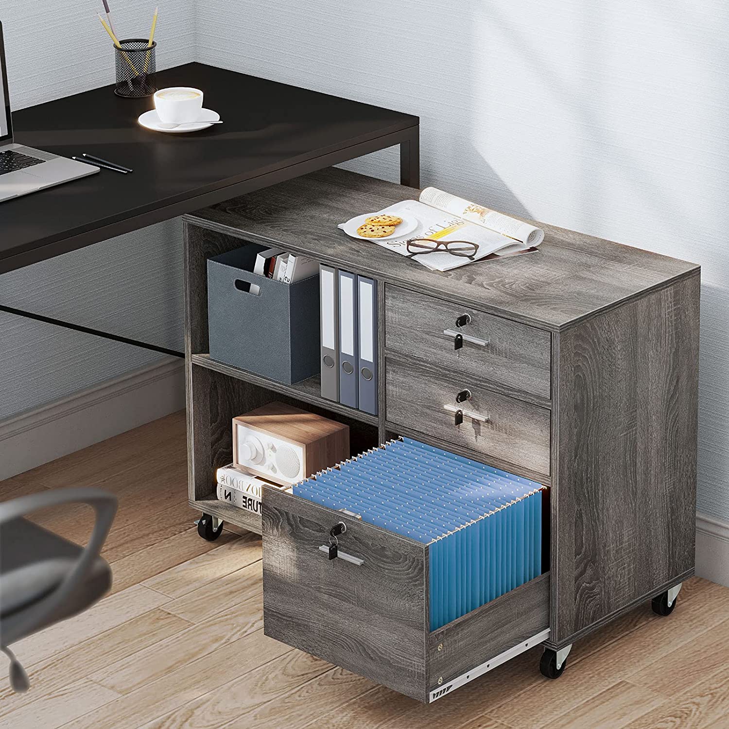 A gray wood-toned mobile filing cabinet with open shelves and drawers sits on a light wood floor under a black office desk.