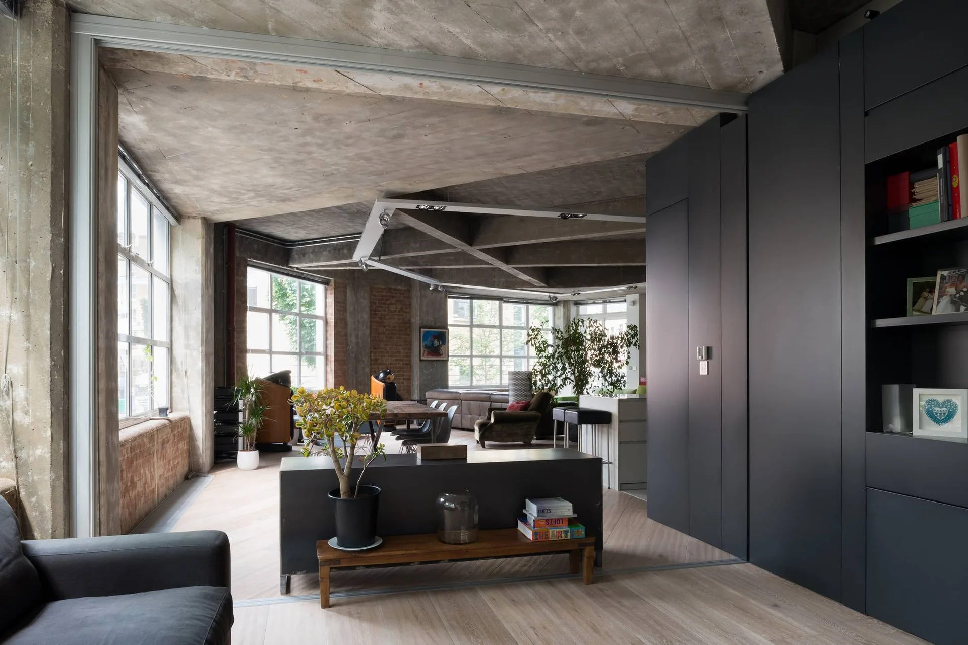 An industrial loft living room with exposed concrete ceilings, a large dark grey cabinet unit, herringbone wood flooring, and large windows.