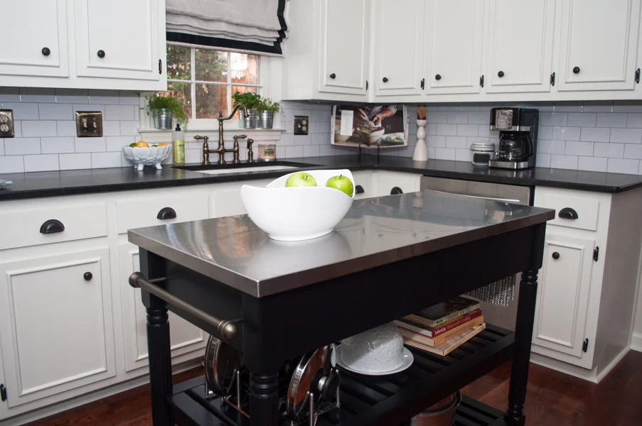 A kitchen with white shaker cabinets, dark countertops, a white subway tile backsplash, and a black kitchen island with a stainless steel top.