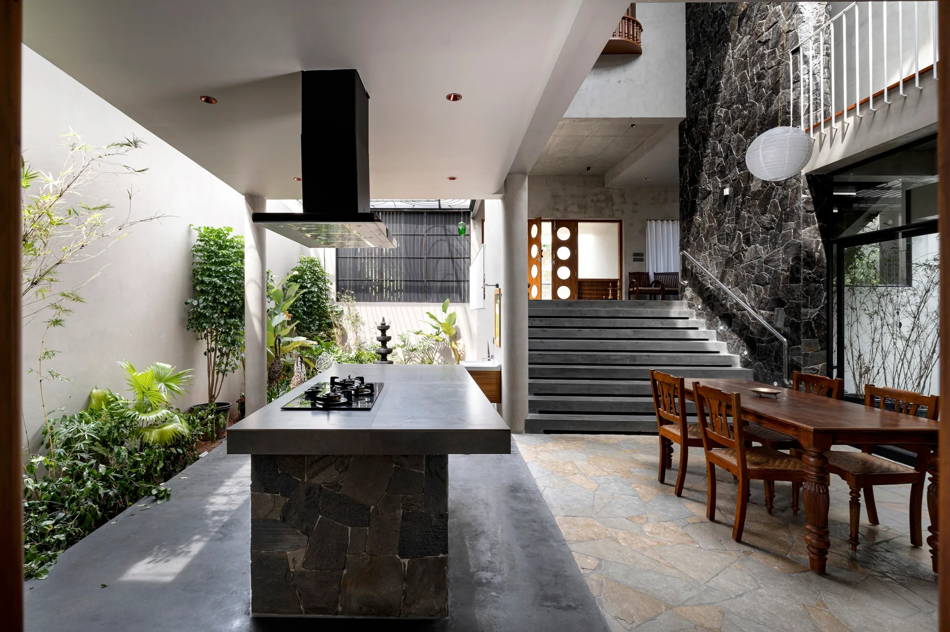 An open atrium kitchen featuring a stone clad island, a wooden dining table with chairs, a concrete staircase, and a dark stone accent wall.
