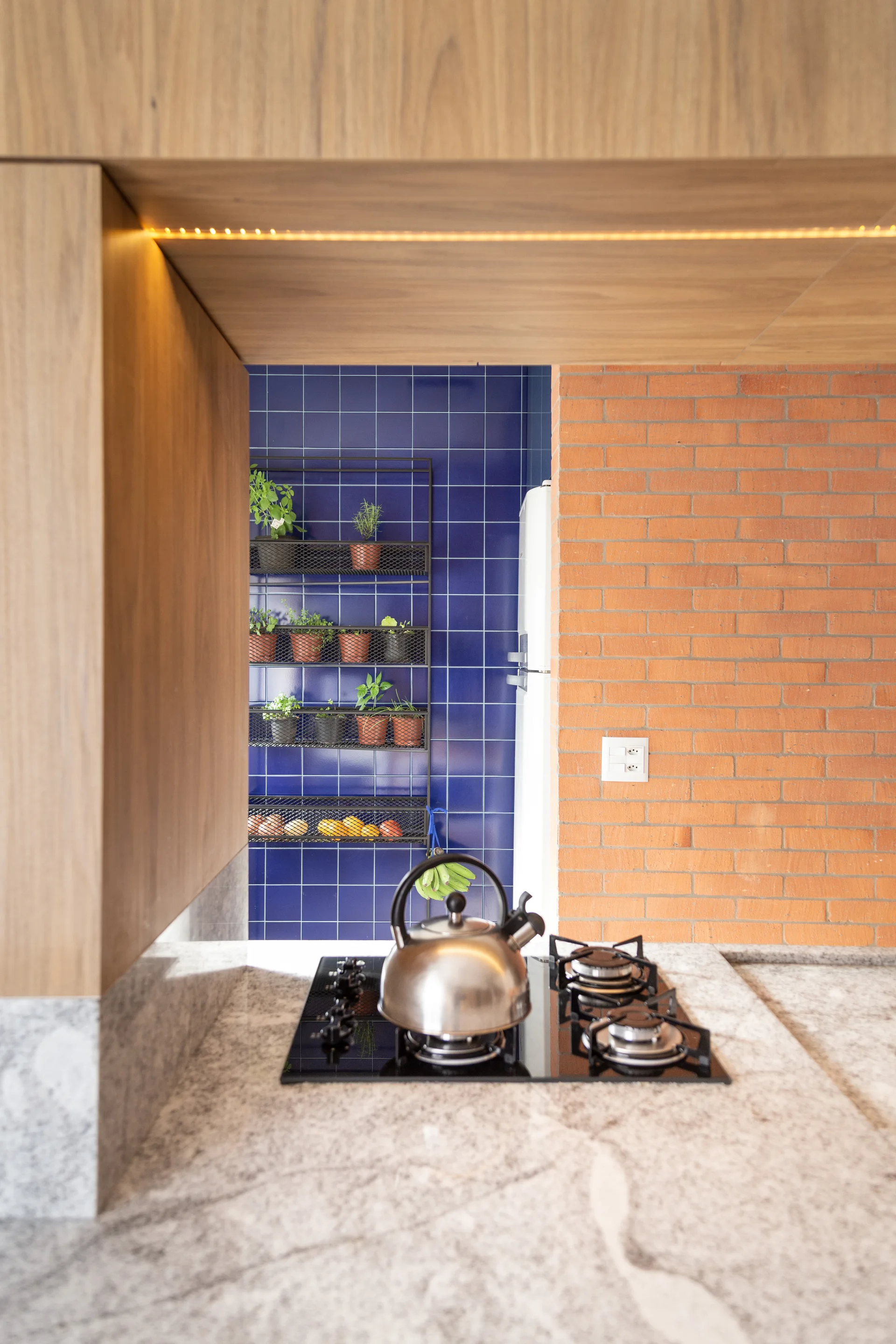 A kitchen stove on a gray stone countertop with a stainless steel kettle, set against a blue tiled wall and an exposed brick wall.