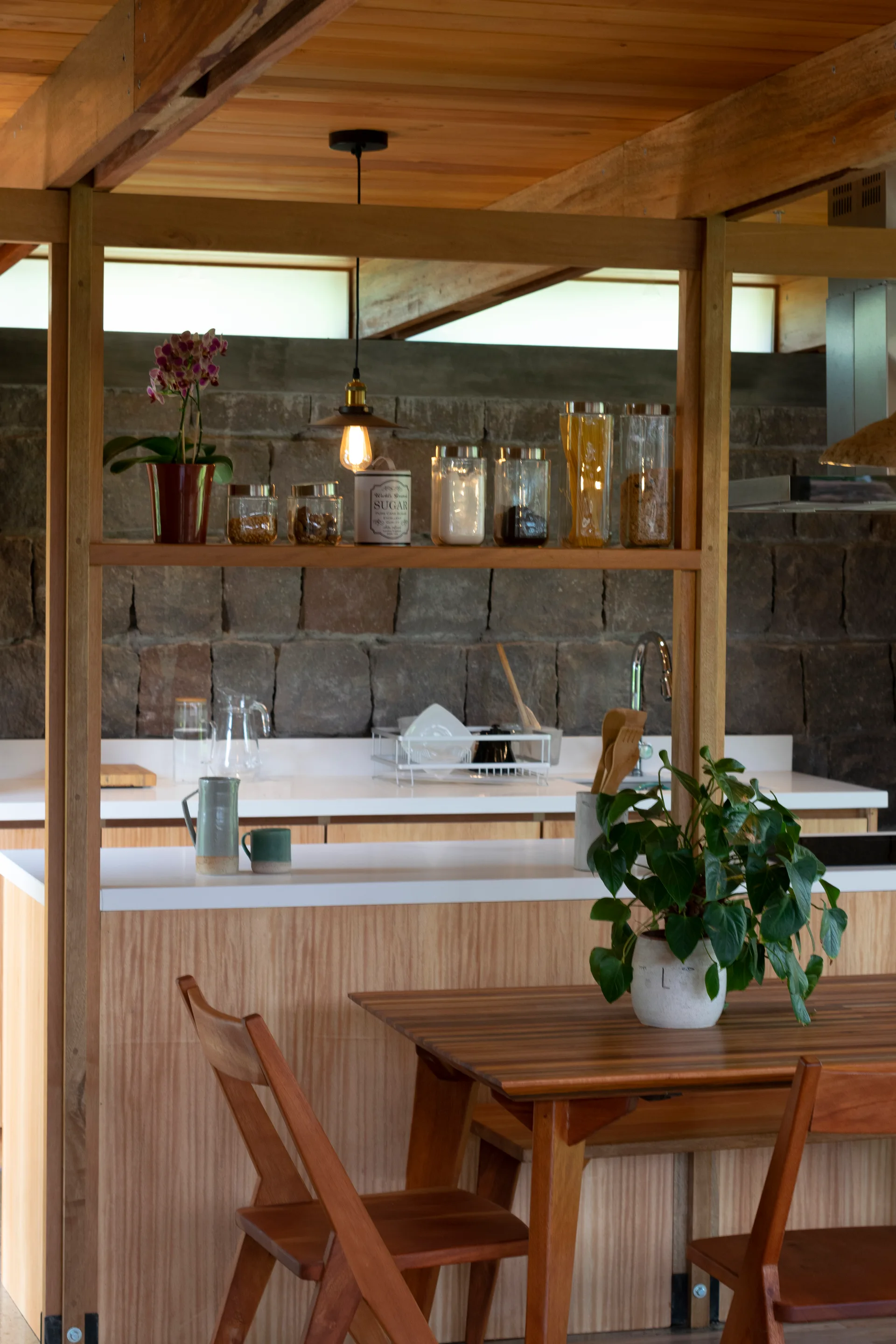 A kitchen featuring a natural wood island and ceiling, a stone masonry wall background, and a wooden dining table with a potted plant.