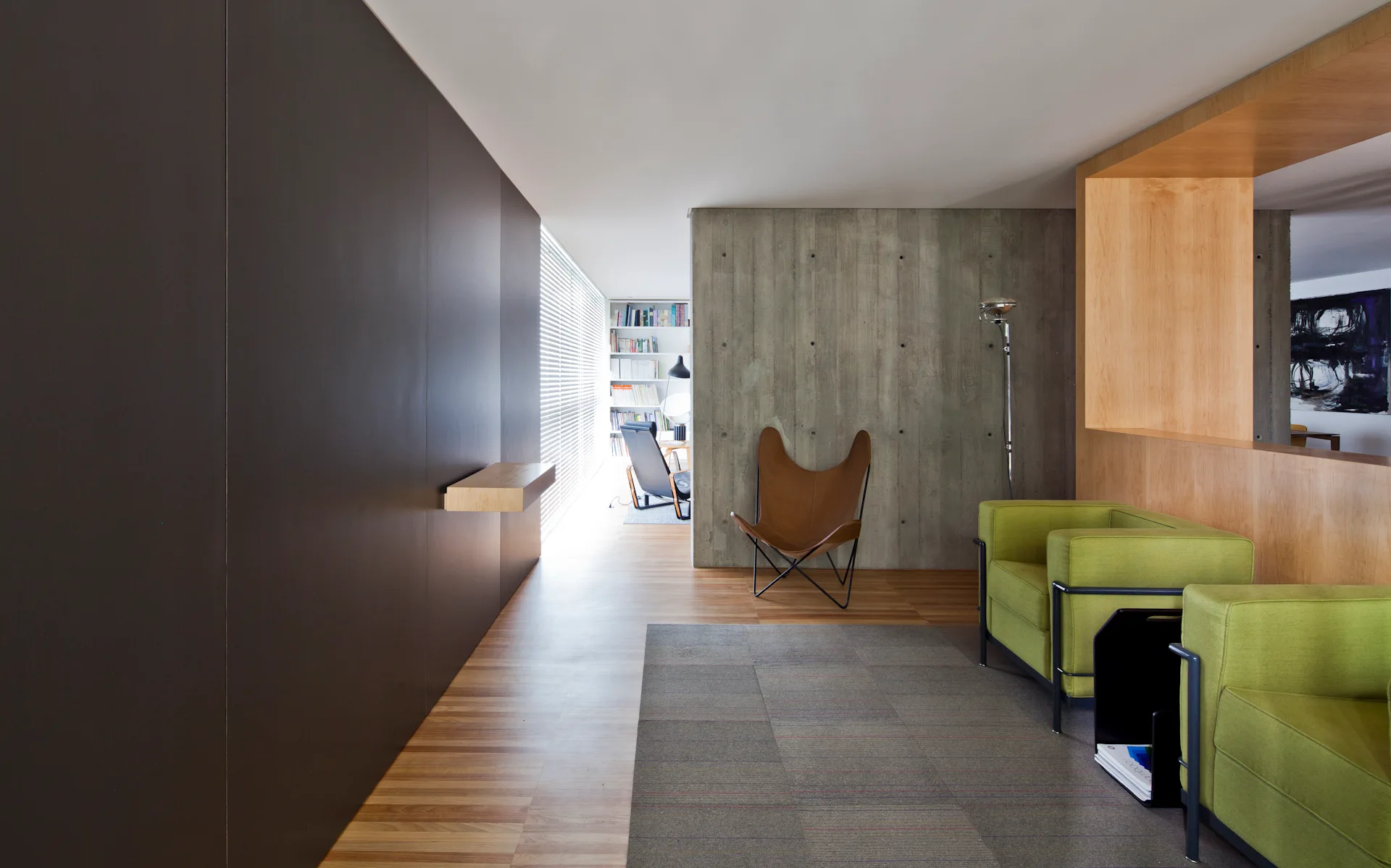 A modern minimalist living room featuring an exposed concrete wall, a tan leather butterfly chair, and two olive green armchairs.