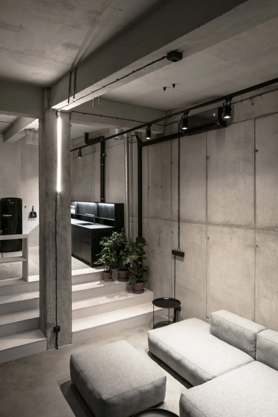 A modern split-level loft living room features a grey modular sofa, exposed concrete walls, and steps leading to a dark kitchen area.