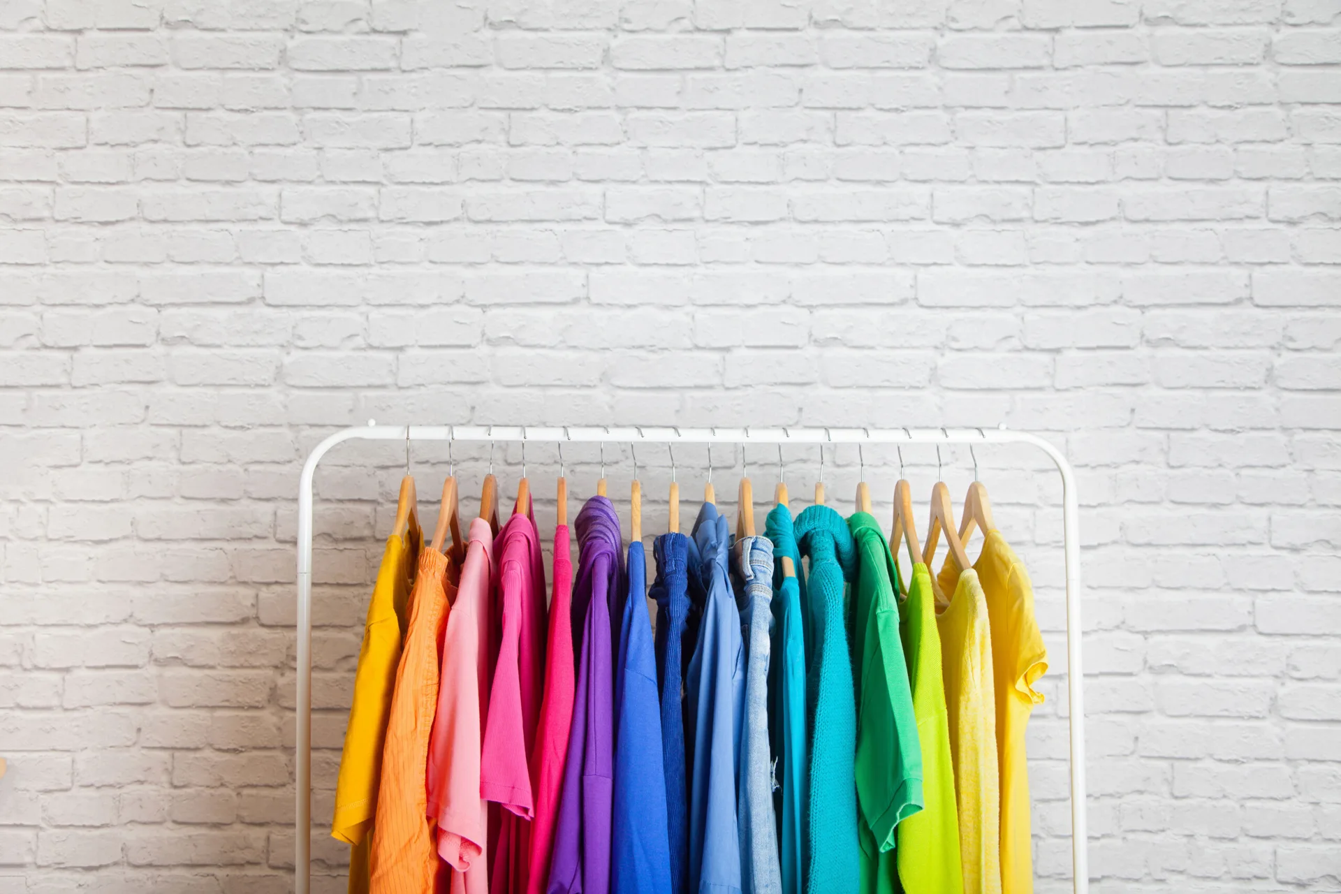 A white metal clothing rack displays colorful garments arranged in rainbow order on wooden hangers against a white brick wall.