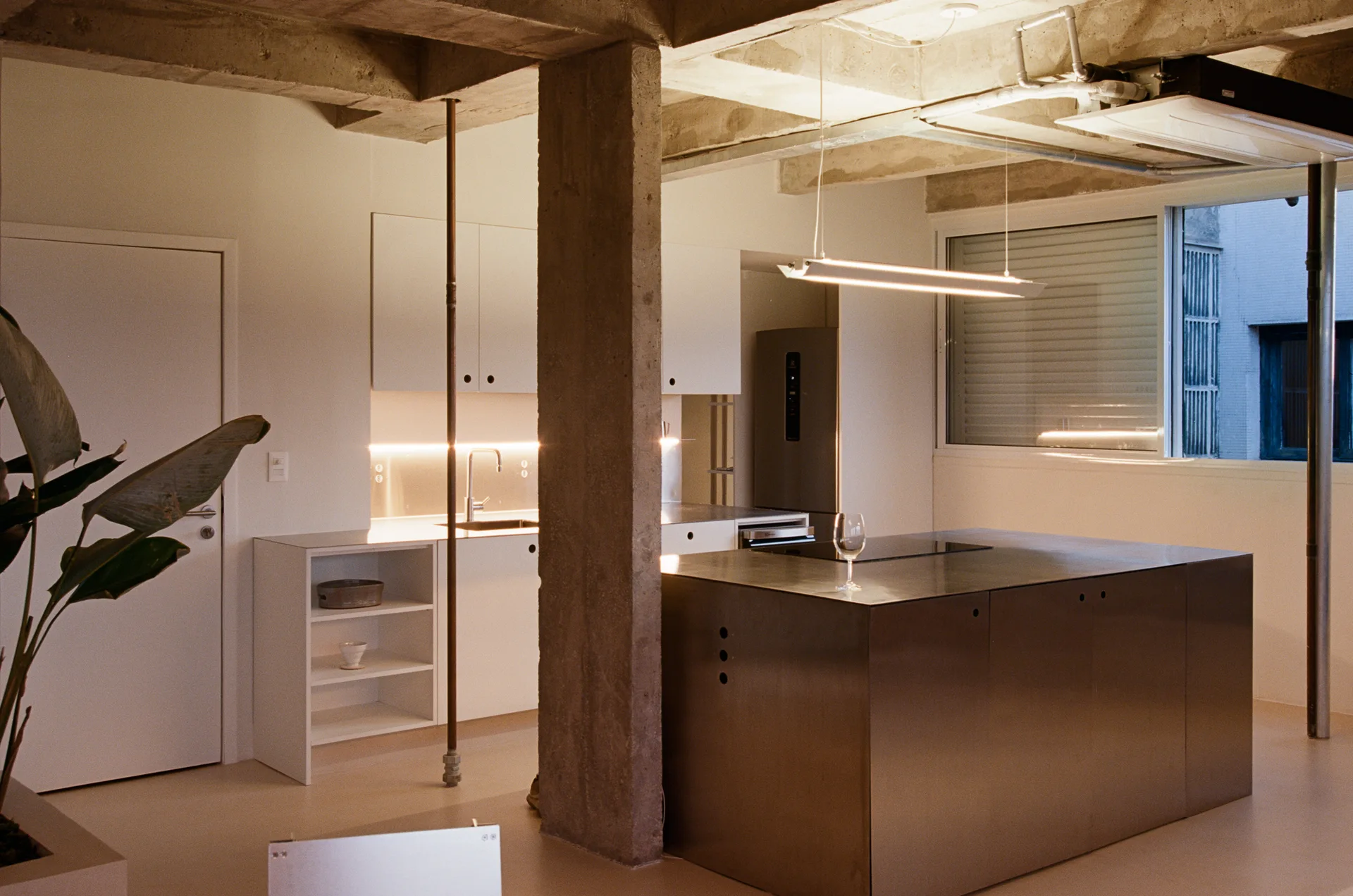 A minimalist industrial kitchen features a large stainless steel island, white cabinetry, a rough concrete pillar, and an exposed concrete ceiling.