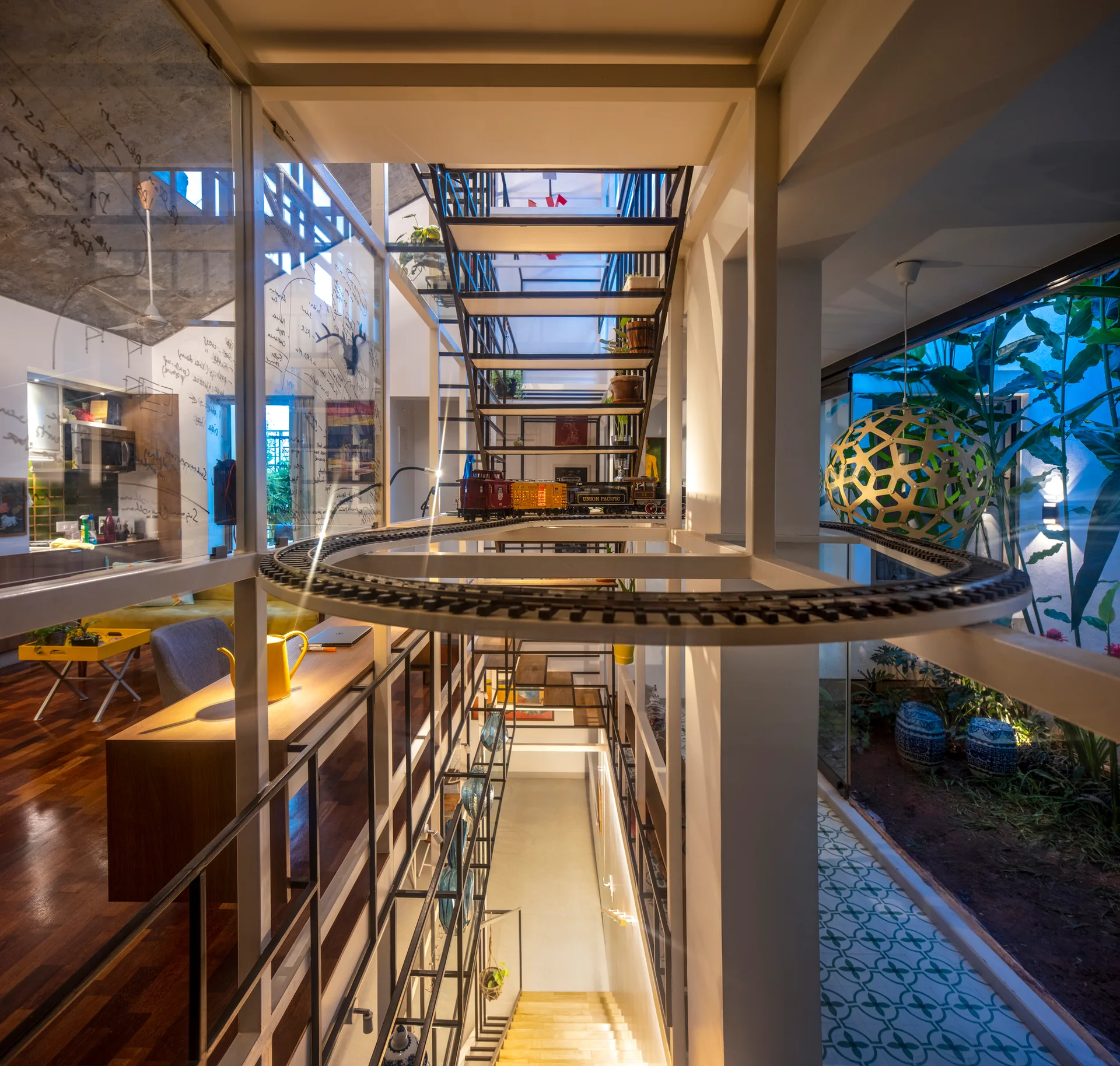Looking down an open metal staircase with a toy train set on a landing, a handwritten glass partition, and an indoor garden below.