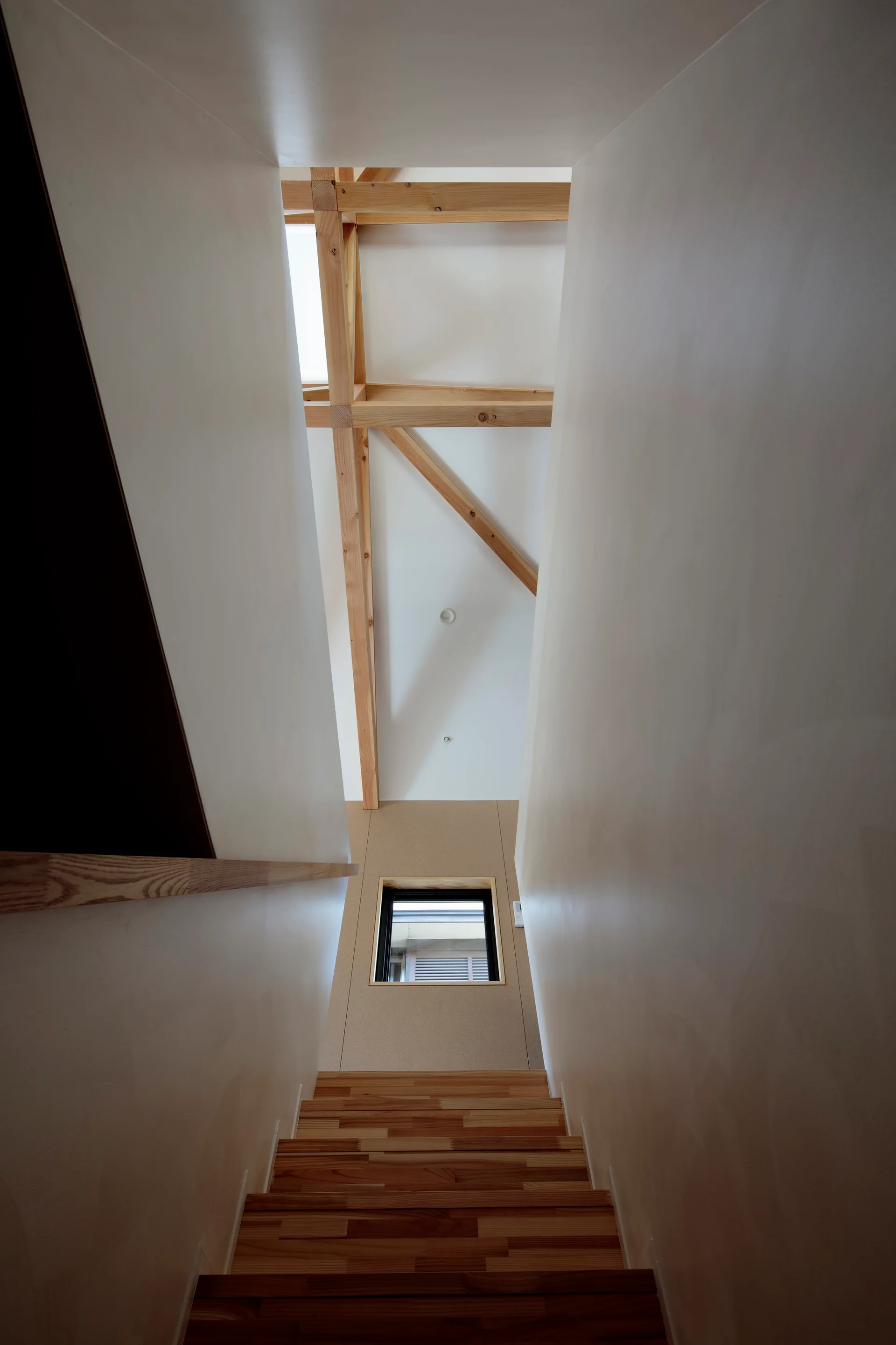 Looking up a narrow wooden staircase framed by smooth white walls toward exposed timber ceiling beams and a skylight.