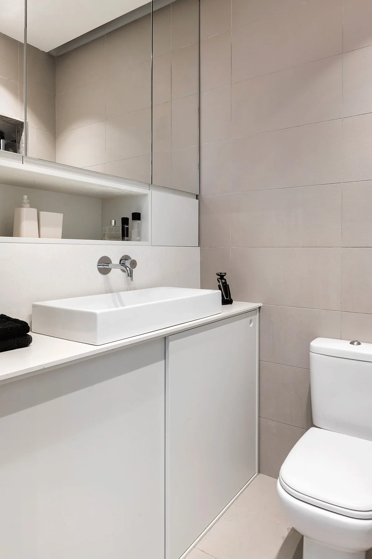 A minimalist bathroom features a white flat-front vanity with a vessel sink, a wall-mounted chrome faucet, and large-format beige wall tiles.