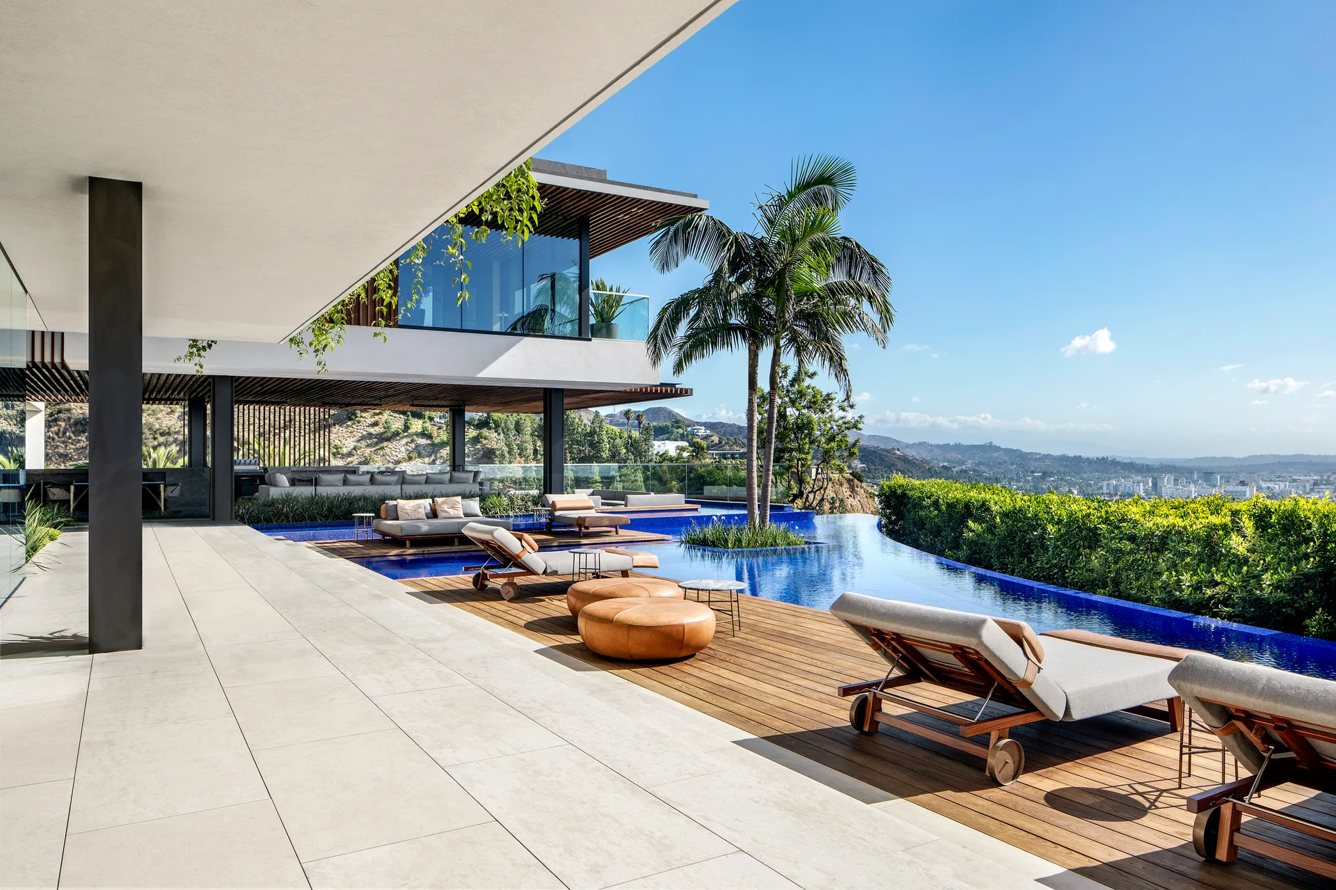 A modern concrete and wood patio features a blue infinity pool, lounge chairs, leather ottomans, and a hillside city view under a clear sky.