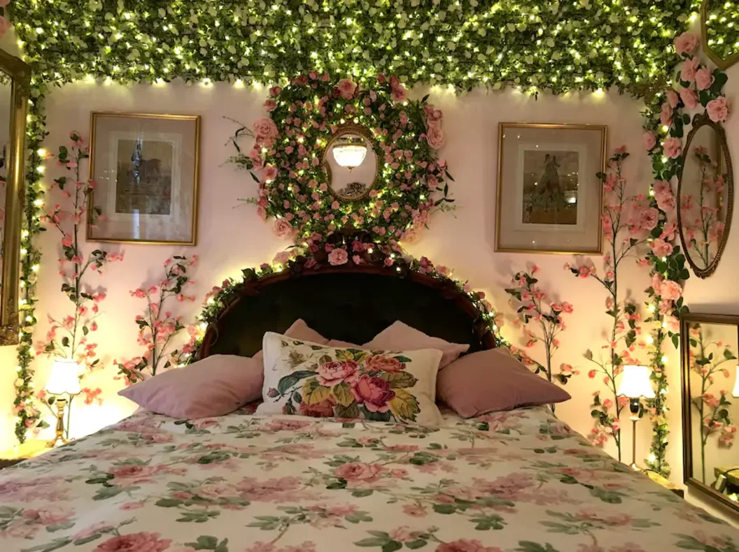 A bedroom with a floral pattern bed, featuring walls decorated with pink artificial flower garlands, green vines, warm fairy lights, and mirrors.