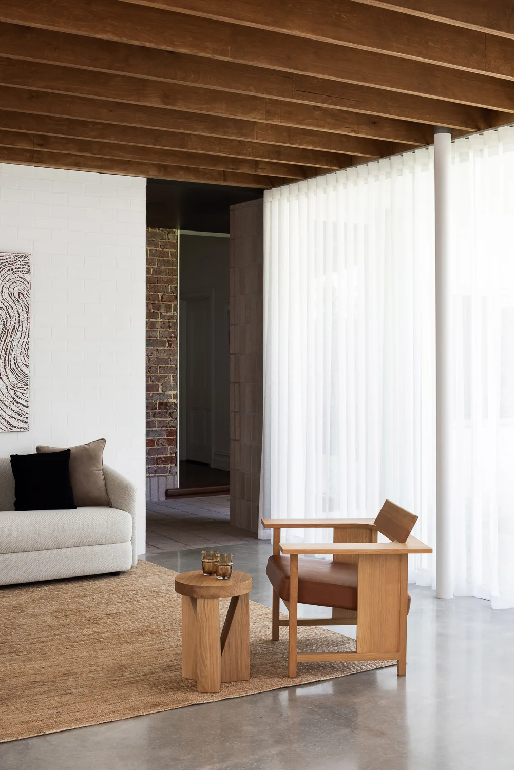 A living room with exposed wood ceiling beams, white painted brick walls, a wooden armchair, a beige sofa, and sheer white floor-to-ceiling curtains.