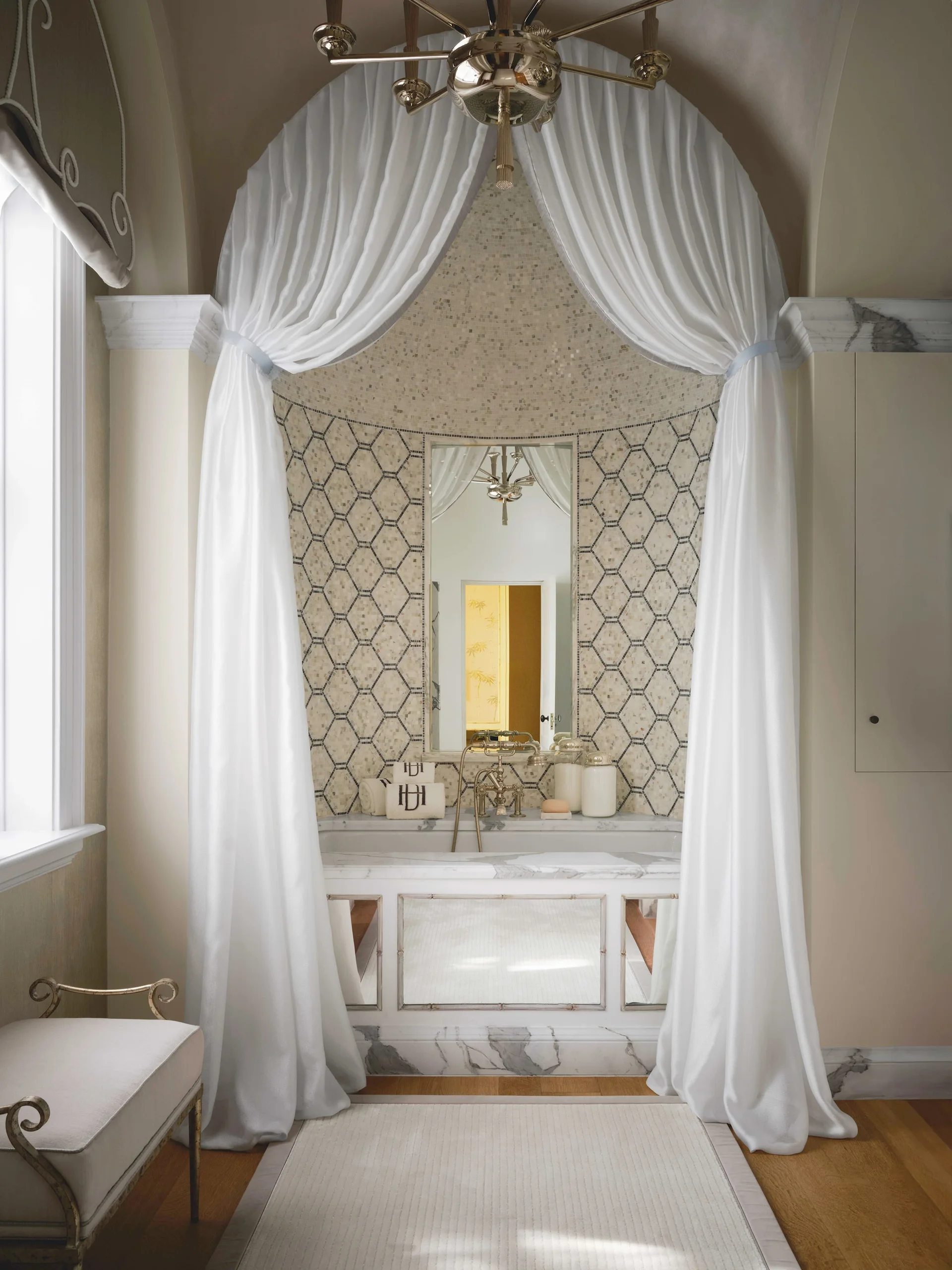 A bathroom alcove featuring a built-in marble bathtub flanked by draped white curtains beneath a curved ceiling with hexagonal mosaic wall tiles.
